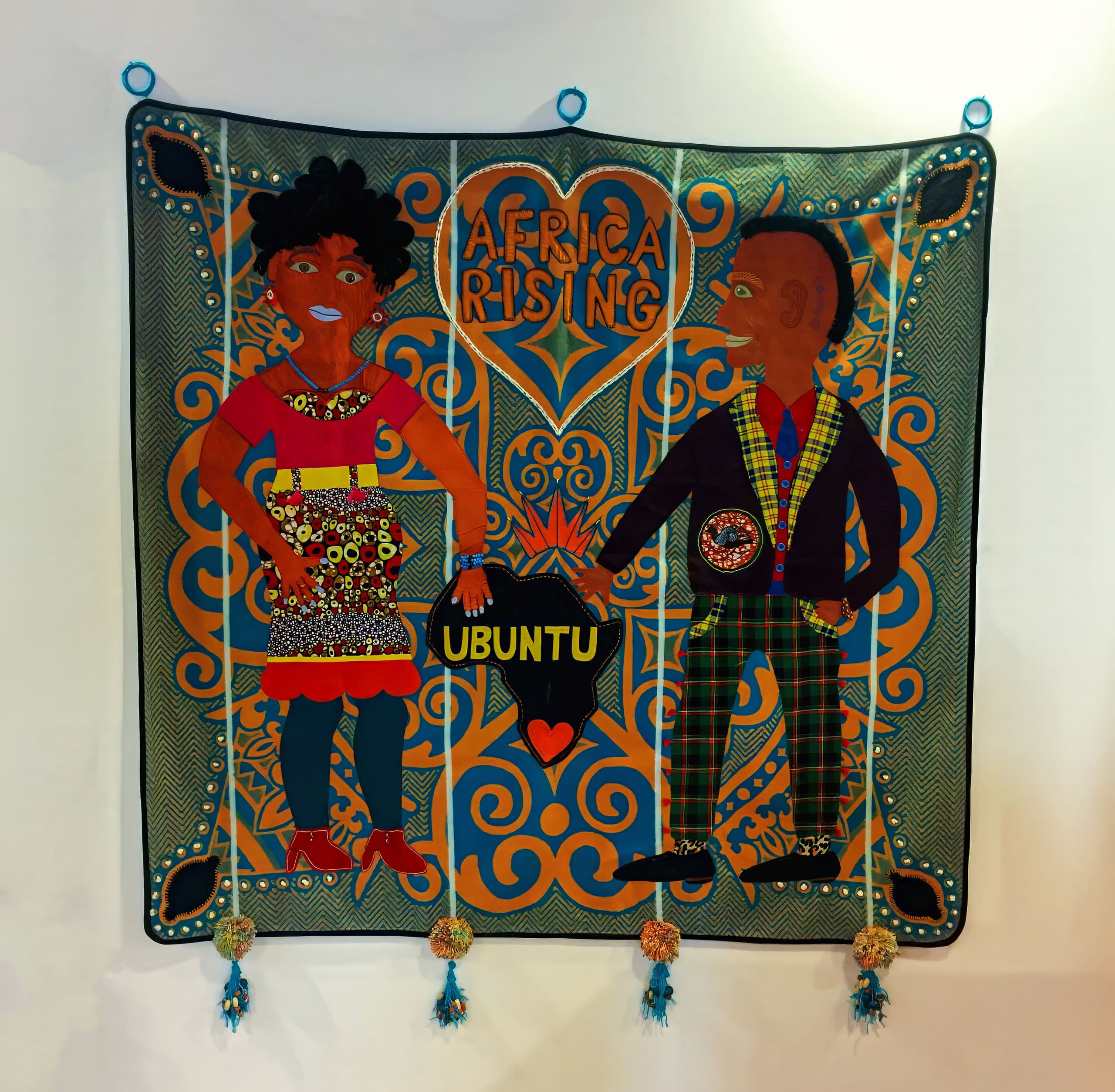 A colourful textile artwork shows two people in vibrant clothing holding hands. Above them is the text “AFRICA RISING.” Between them, they hold a heart and a sign reading “UBUNTU.” The background has intricate orange and blue patterns with four tassels at the bottom edge.