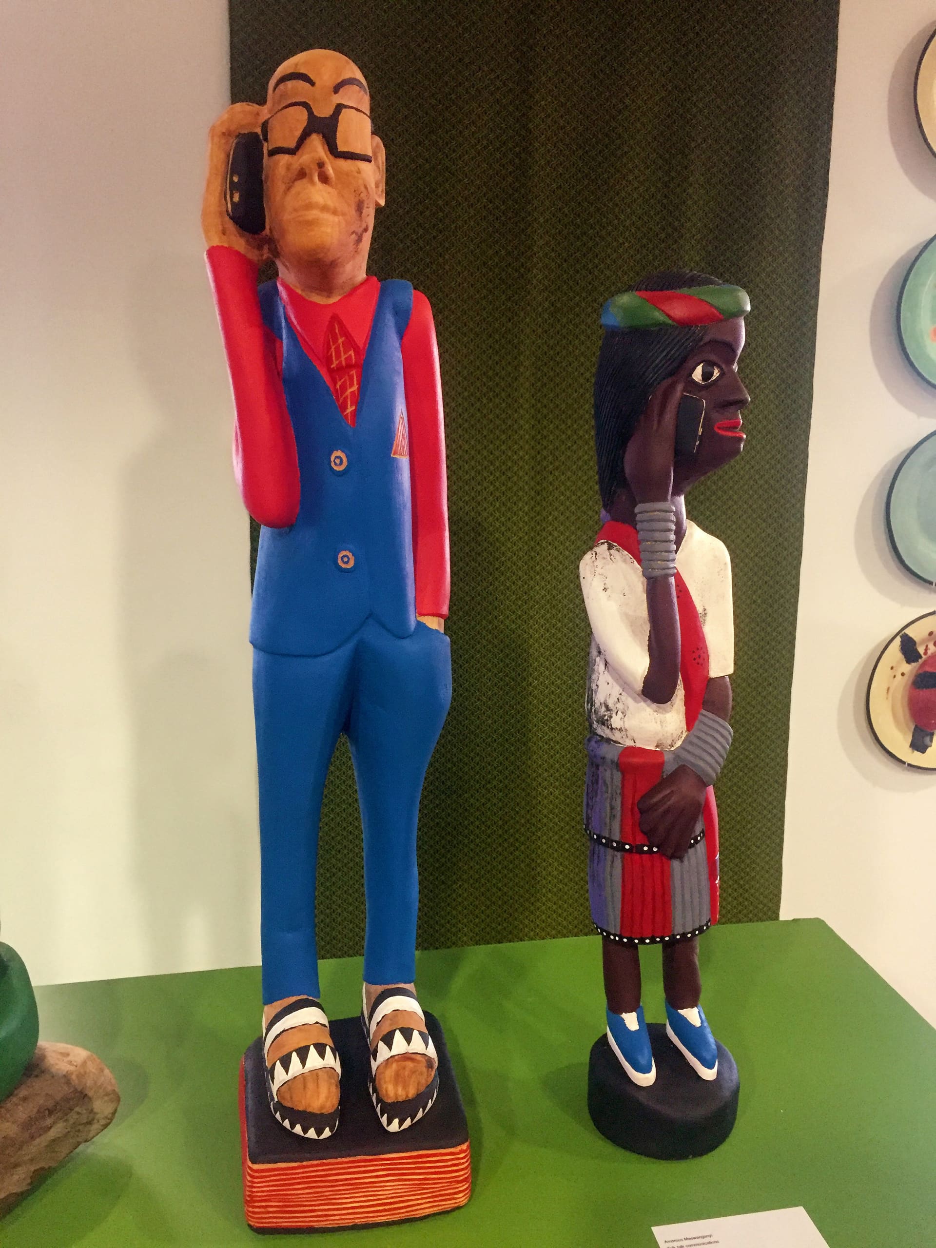 Two colourful wooden sculptures stand on a green surface. The left figure is a man in a blue suit and red tie, wearing glasses, holding a phone to his ear. The right figure is a woman in traditional dress, also holding a phone to her ear. Both are detailed and vibrant.