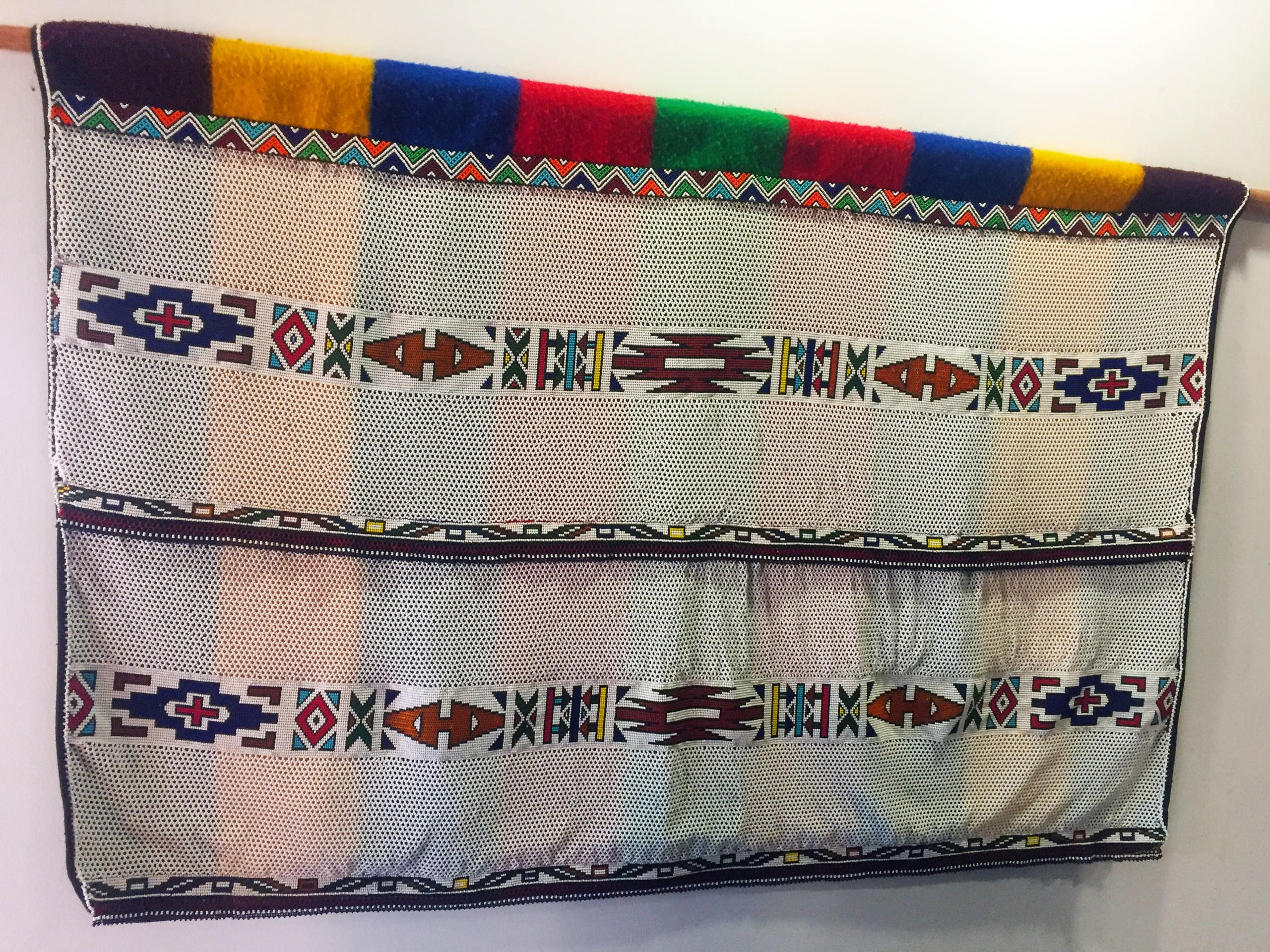 A colourful Basotho blanket hangs on a wall. It features geometric patterns with earthy tones, including brown, blue, white, and red, bordered by a row of vibrant coloured squares at the top. The fabric has a textured, woven appearance.