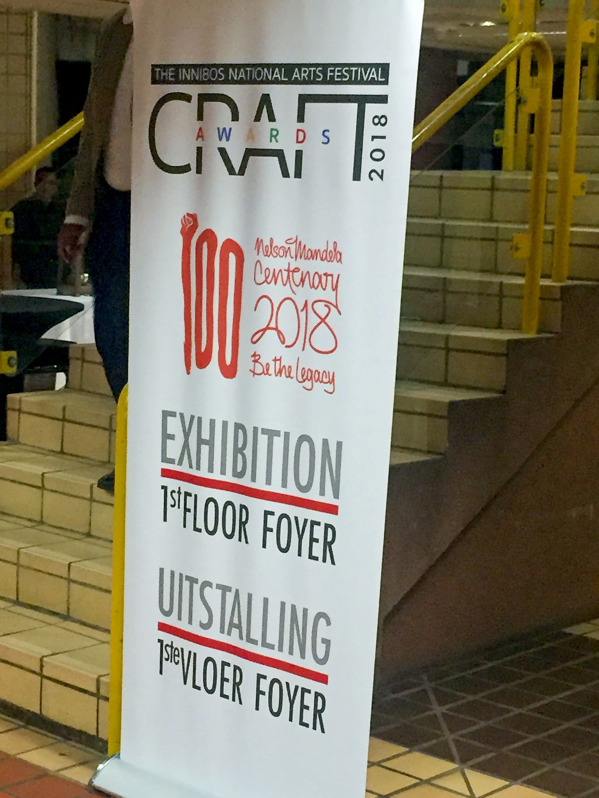 A vertical banner displays “CRAFT AWARDS 2018” and “Nelson Mandela Centenary 2018 Be the legacy.” It advertises an exhibition on the first floor foyer in both English and Afrikaans: “1st FLOOR FOYER” and “1STE VLOER FOYER.” The banner stands by tiled stairs with yellow rails.