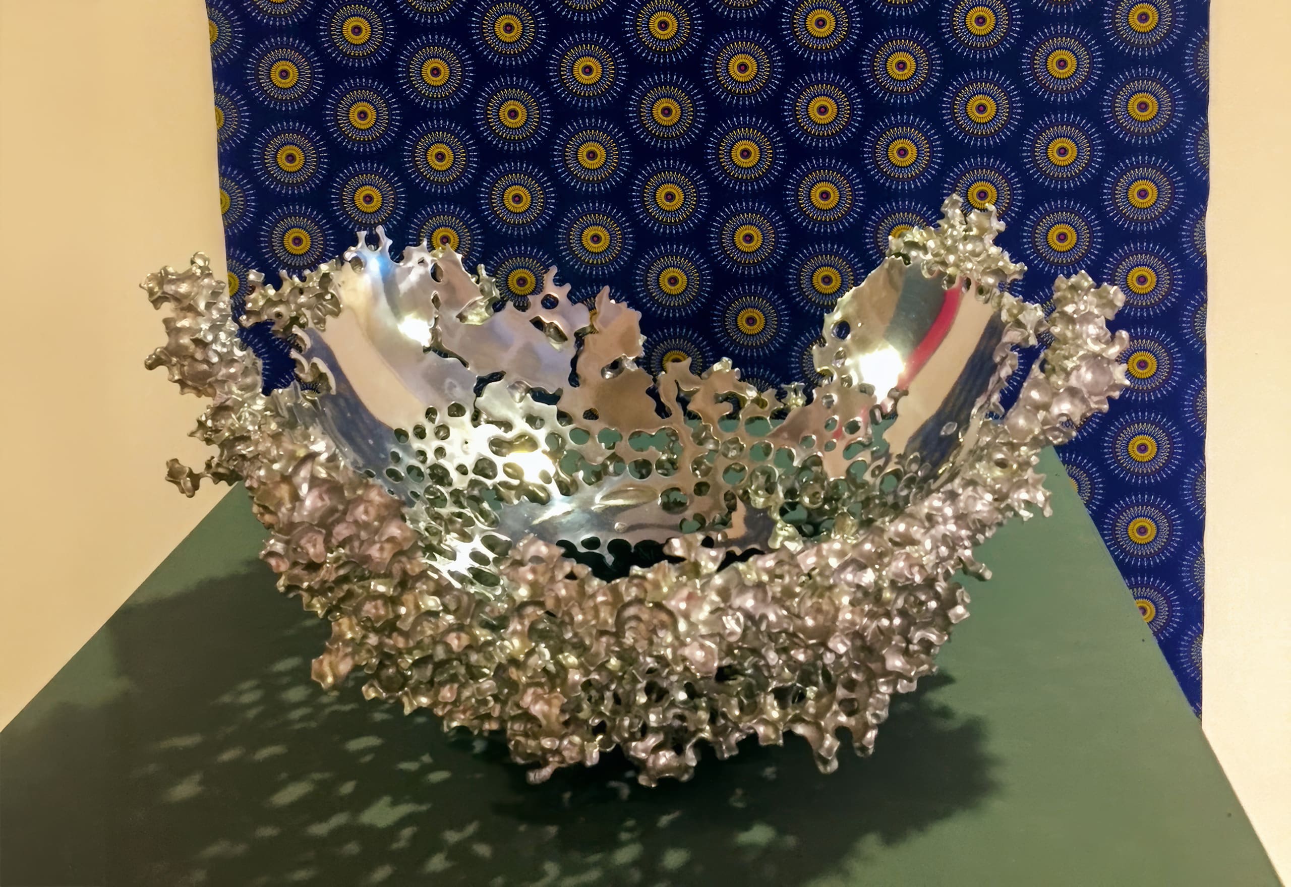 A silver, textured metal bowl with an irregular, jagged rim sits on a green surface. The inner part is smooth and reflective, whilst the outer edge features clustered, organic shapes. Behind it is a blue fabric with a yellow and green circular pattern.