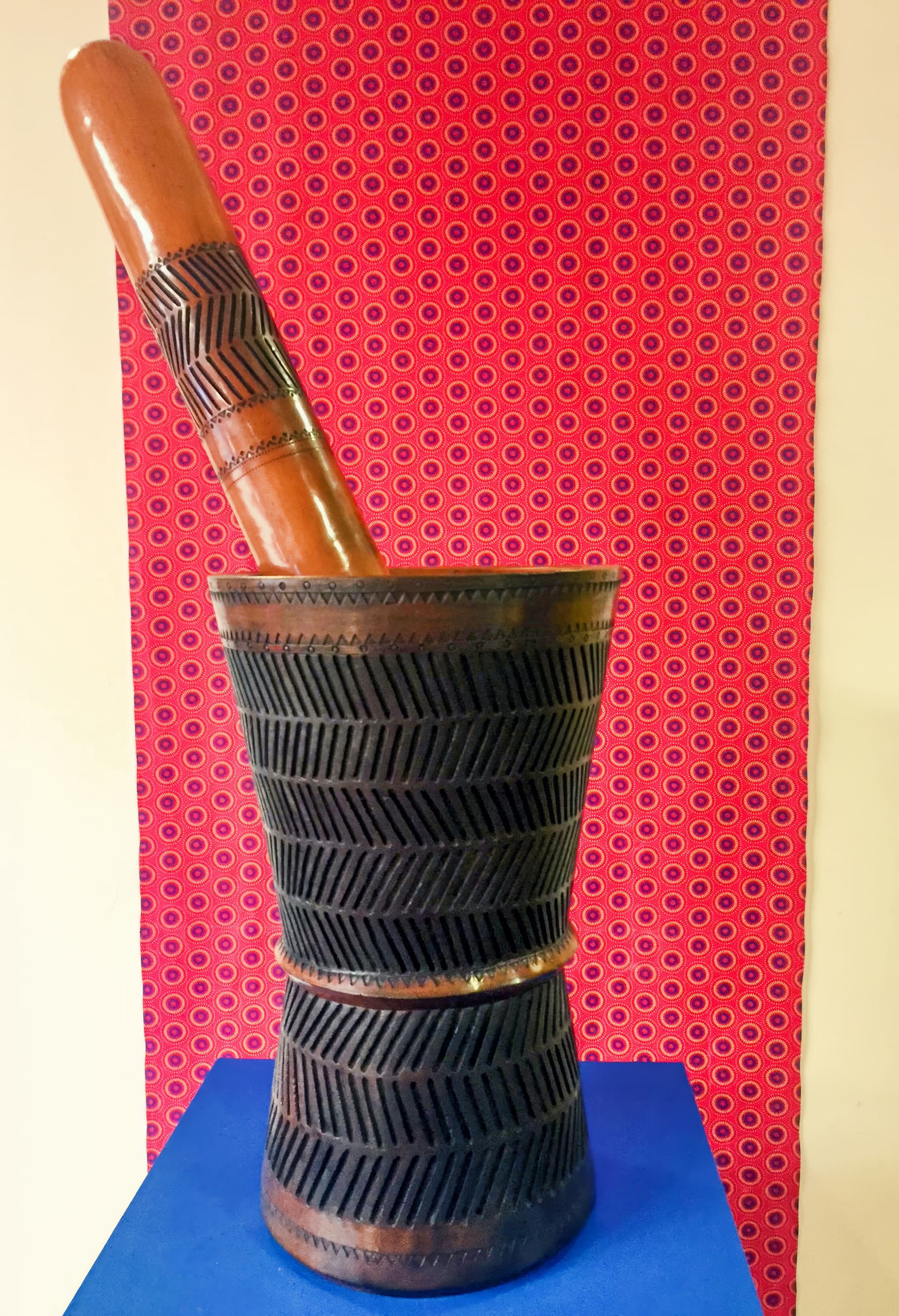 A large, dark wooden mortar with engraved patterns sits on a blue surface. A matching pestle leans inside it. The background is a red fabric with a repetitive circular pattern, bordered by cream-coloured walls.