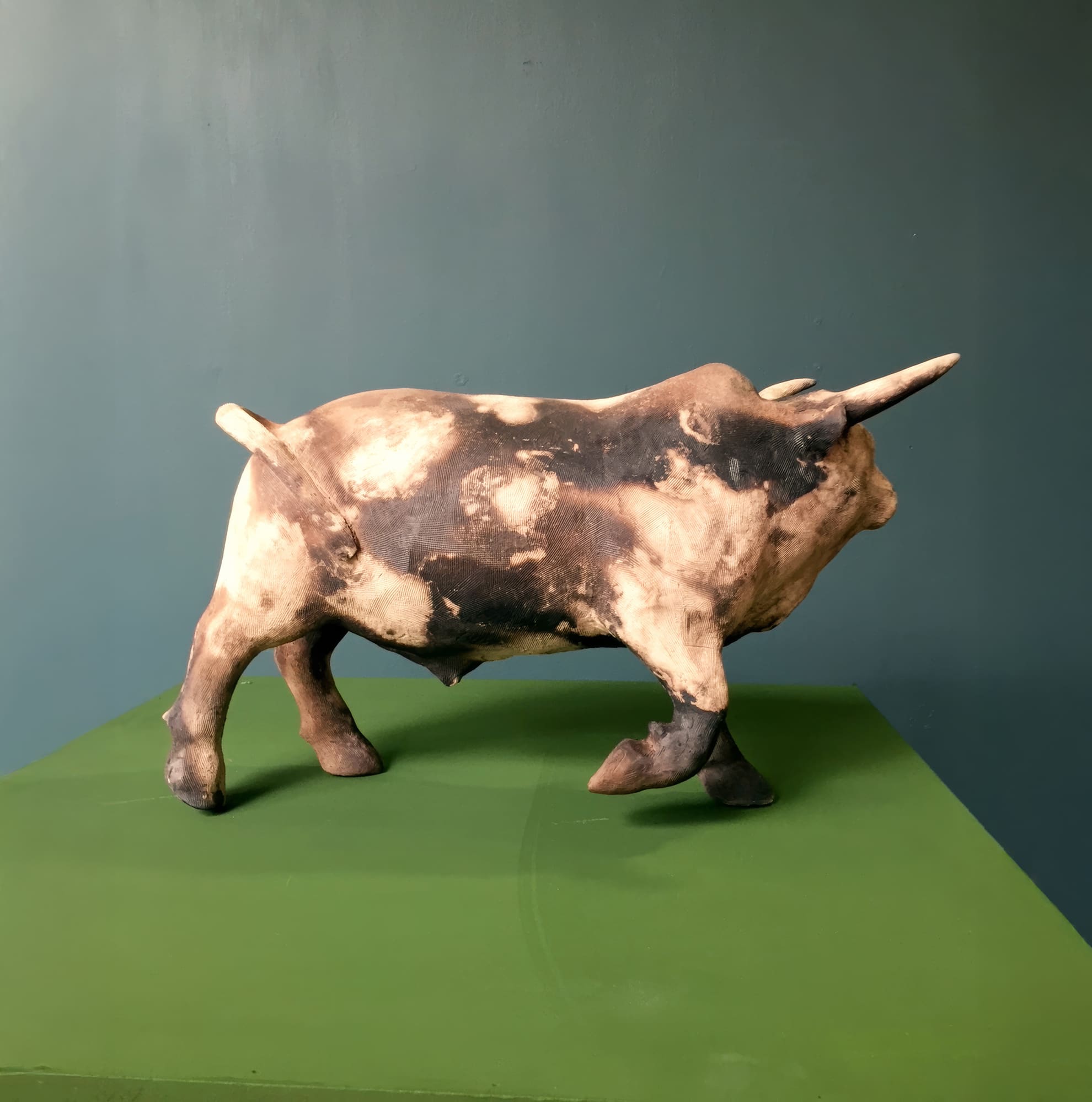 A ceramic bull sculpture with a mottled brown, black, and cream finish stands on a green surface against a plain slate blue wall. The bull faces right, showing its horns, muscular build, and a slightly raised tail.