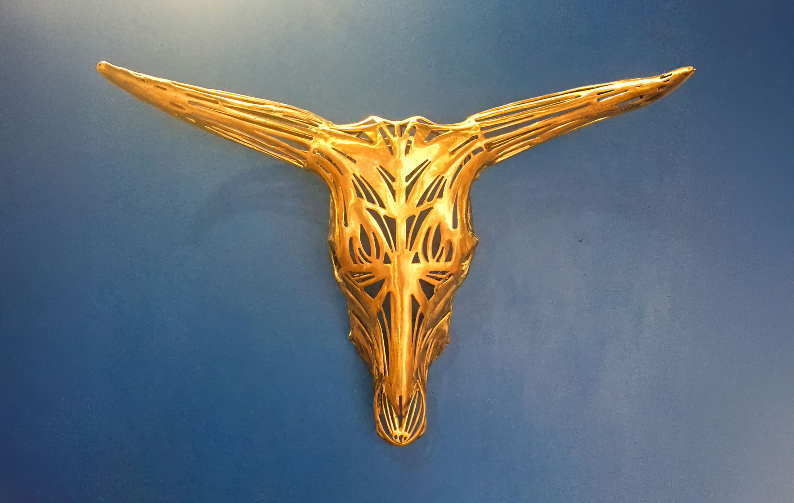 A gold, stylised bull skull sculpture with long, pointed horns is mounted on a smooth blue wall. The skull has an intricate, openwork design with geometric cut-outs, giving it a modern, artistic appearance.