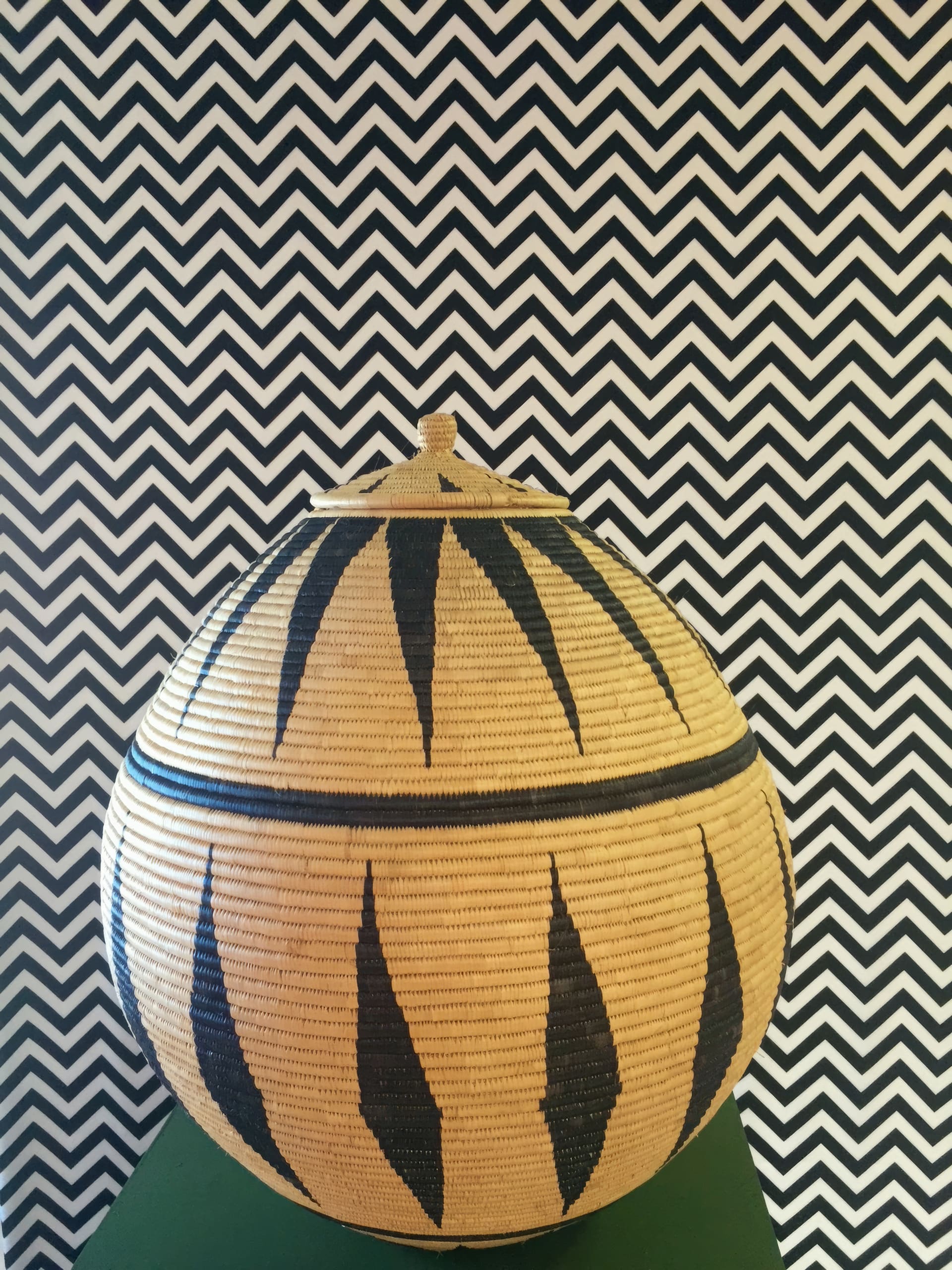A woven, round lidded basket with a geometric black and natural pattern sits on a green surface. The background is a bold black and white zigzag chevron wallpaper, creating a striking contrast with the basket’s design.