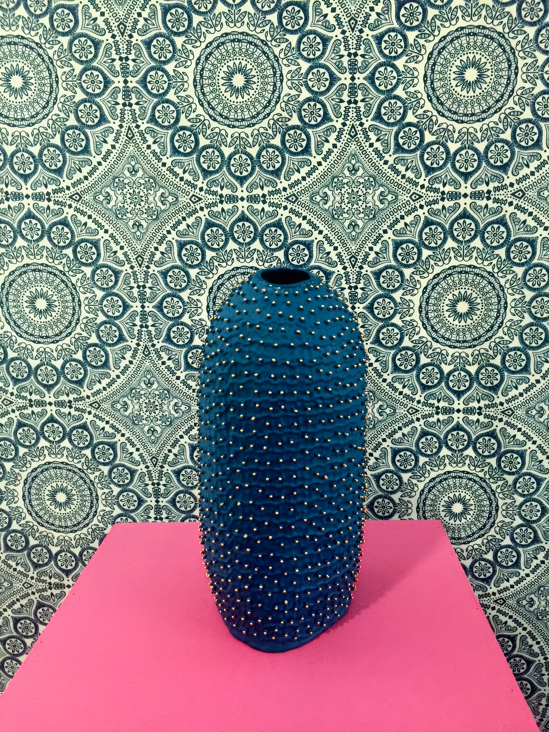 A tall, blue vase with a textured surface covered in small gold studs stands on a bright pink surface. The background features a detailed, circular blue and white geometric patterned wallpaper. The scene is vibrant and visually striking.