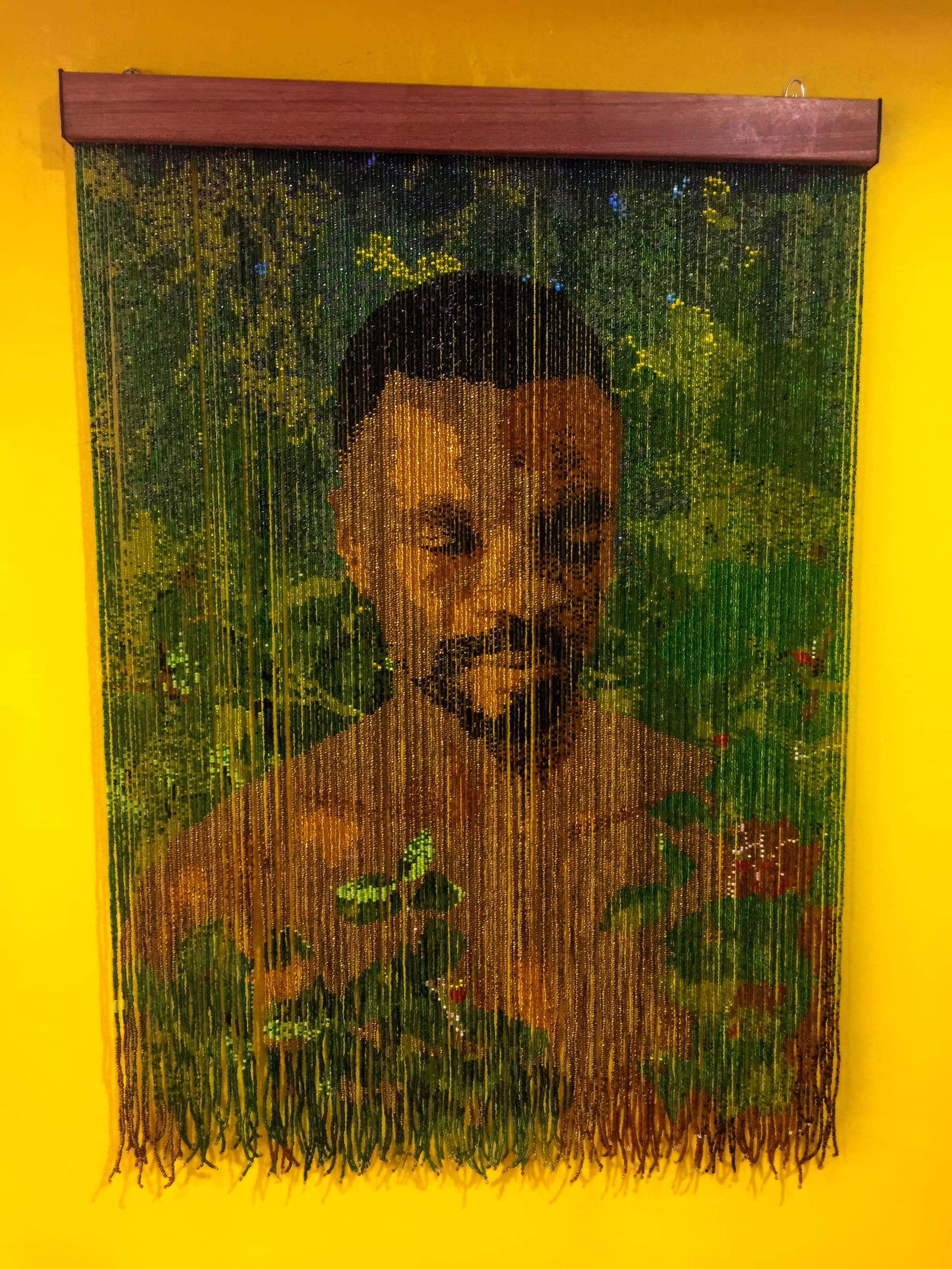 A beaded curtain art piece hangs on a yellow wall. It depicts a man's face and upper body surrounded by green foliage. The beads are arranged to form his features, blending with leafy patterns, creating a layered, semi-transparent portrait effect.