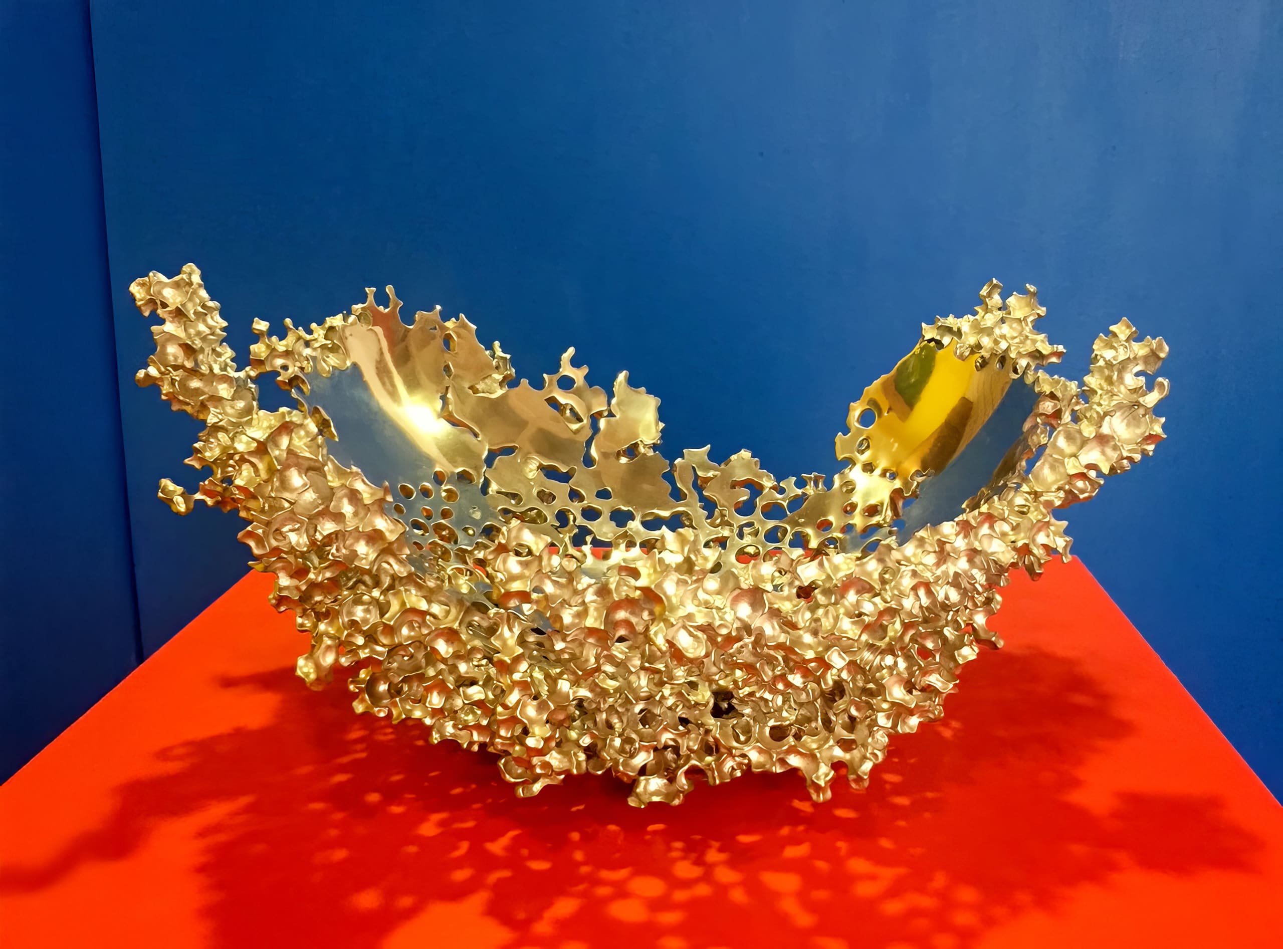 A metallic, abstract bowl with an intricate, irregular lattice design sits on a bright red surface against a solid blue background. The bowl's golden hue and organic, coral-like structure catch and reflect light.