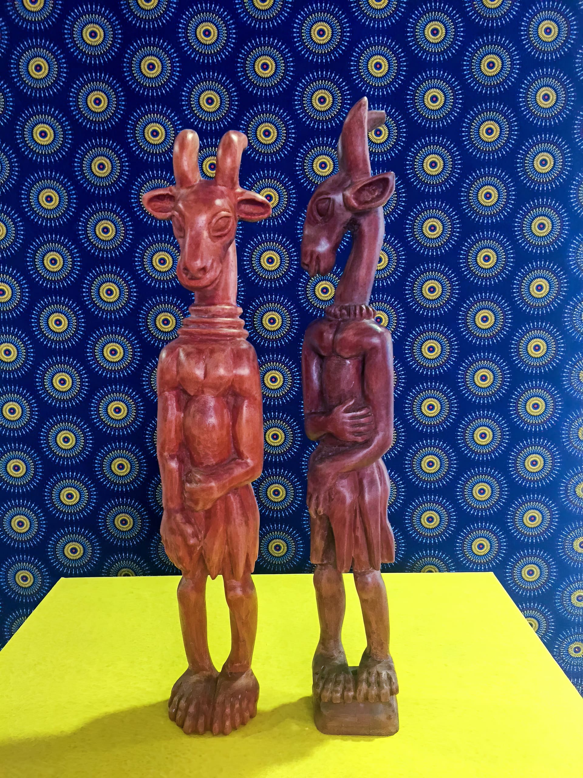 Two carved wooden statues resembling anthropomorphic antelopes stand upright on a bright yellow surface. The background is a blue patterned fabric with concentric yellow and white circles and dots, creating a vibrant and contrasting setting.