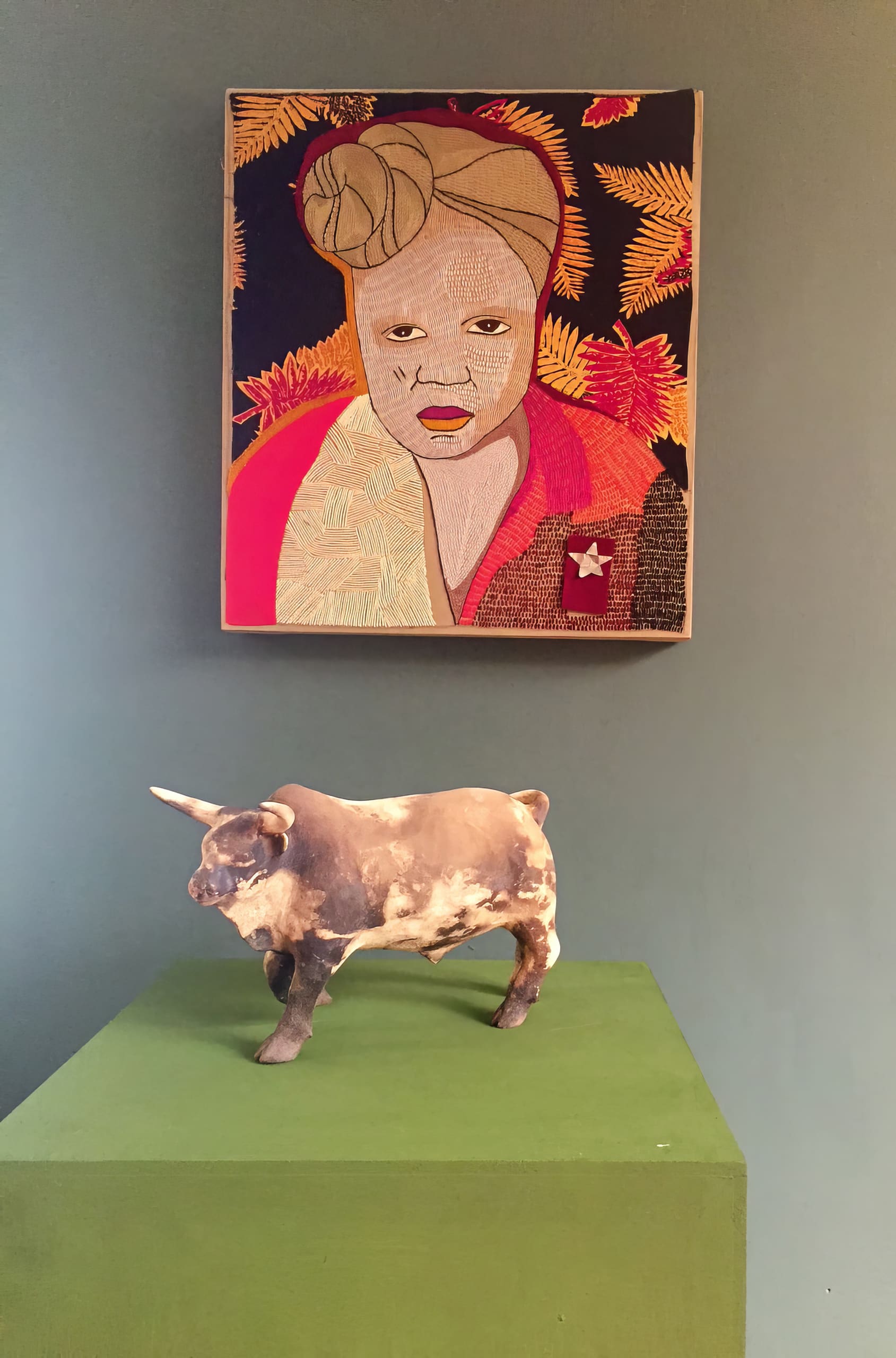 A ceramic bull sculpture stands on a green plinth. Above it hangs a colourful portrait of a person with a headwrap, warm-toned clothes, and a vivid leafy background, set against a muted green wall.