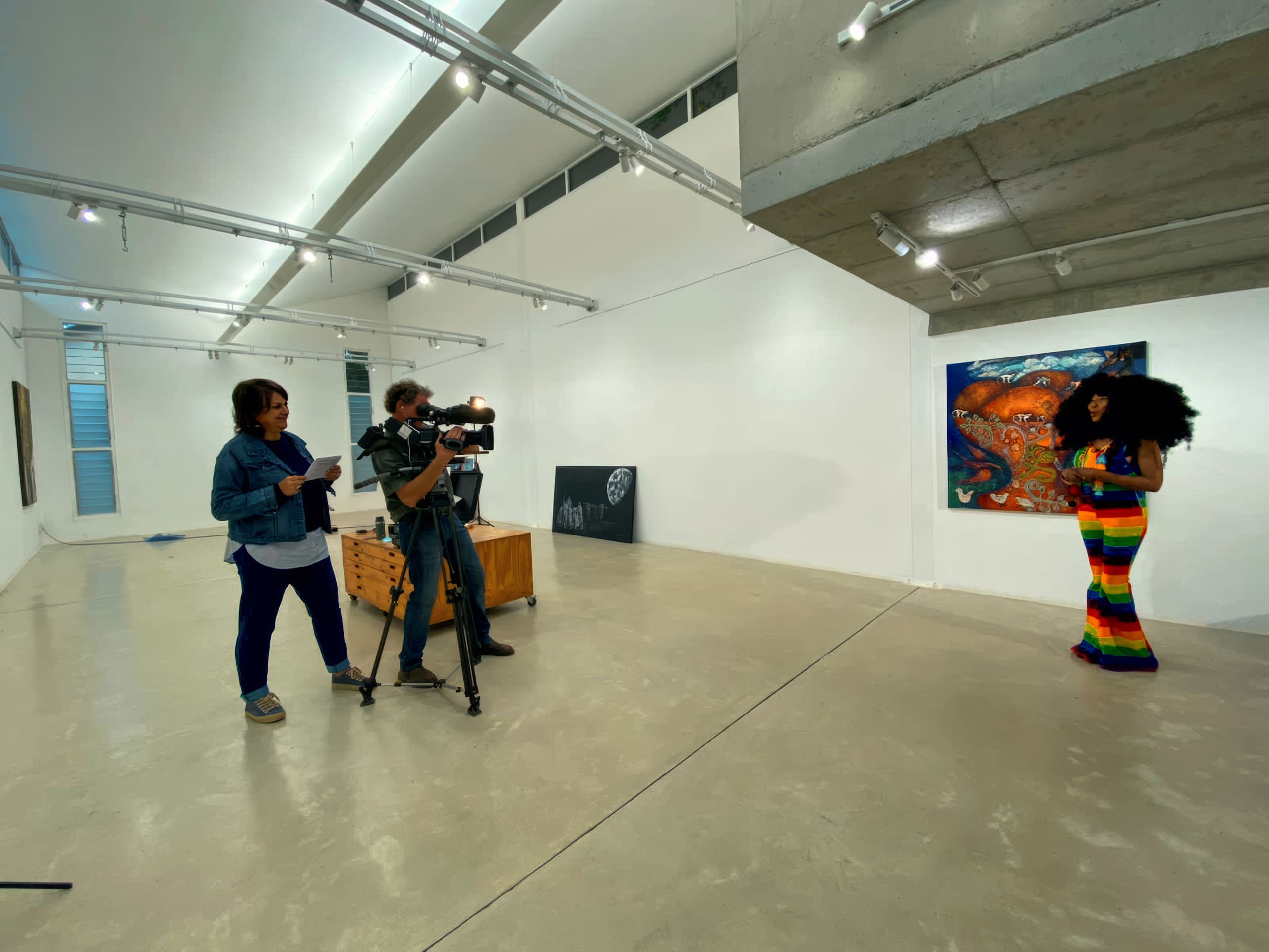 In a modern art gallery, a camera crew films a woman in a colourful rainbow outfit presenting in front of a vivid painting; artworks hang sparsely on white walls, and bright lights illuminate the open, polished concrete space.
