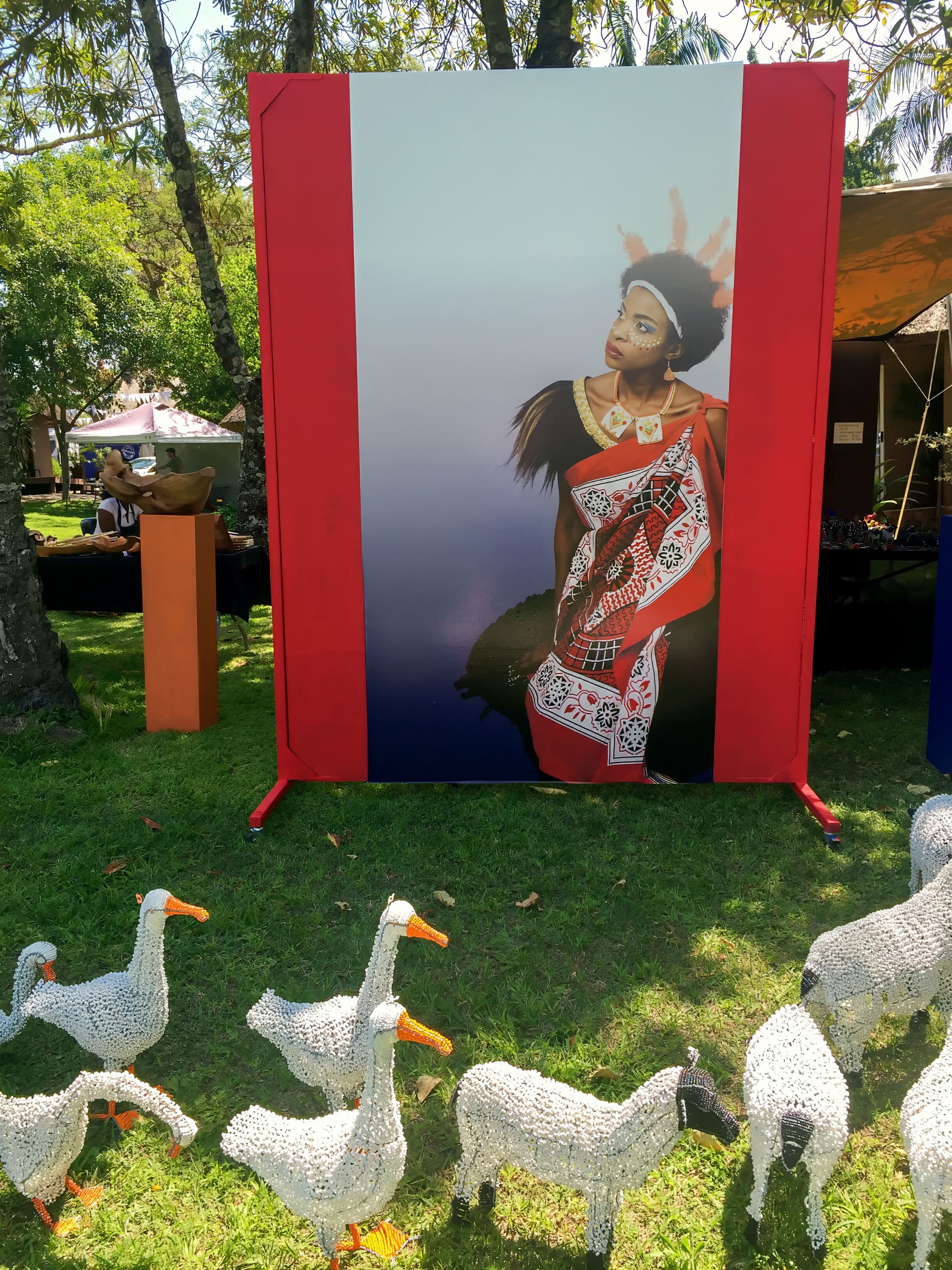 A large photo display shows a woman in traditional attire and a feathered headpiece. In front are white woven sculptures of geese and sheep on green grass. The background includes trees, market stalls, and sunlit outdoor surroundings.