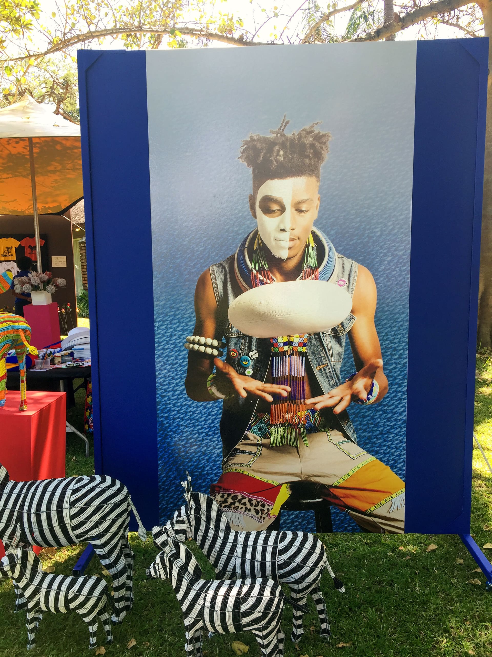A large poster shows a young Black person with face paint, colourful beaded jewellery, and patterned clothing, holding hands under a floating calabash. In front, striped zebra sculptures stand on grass. Trees and a market stall are visible in the background.