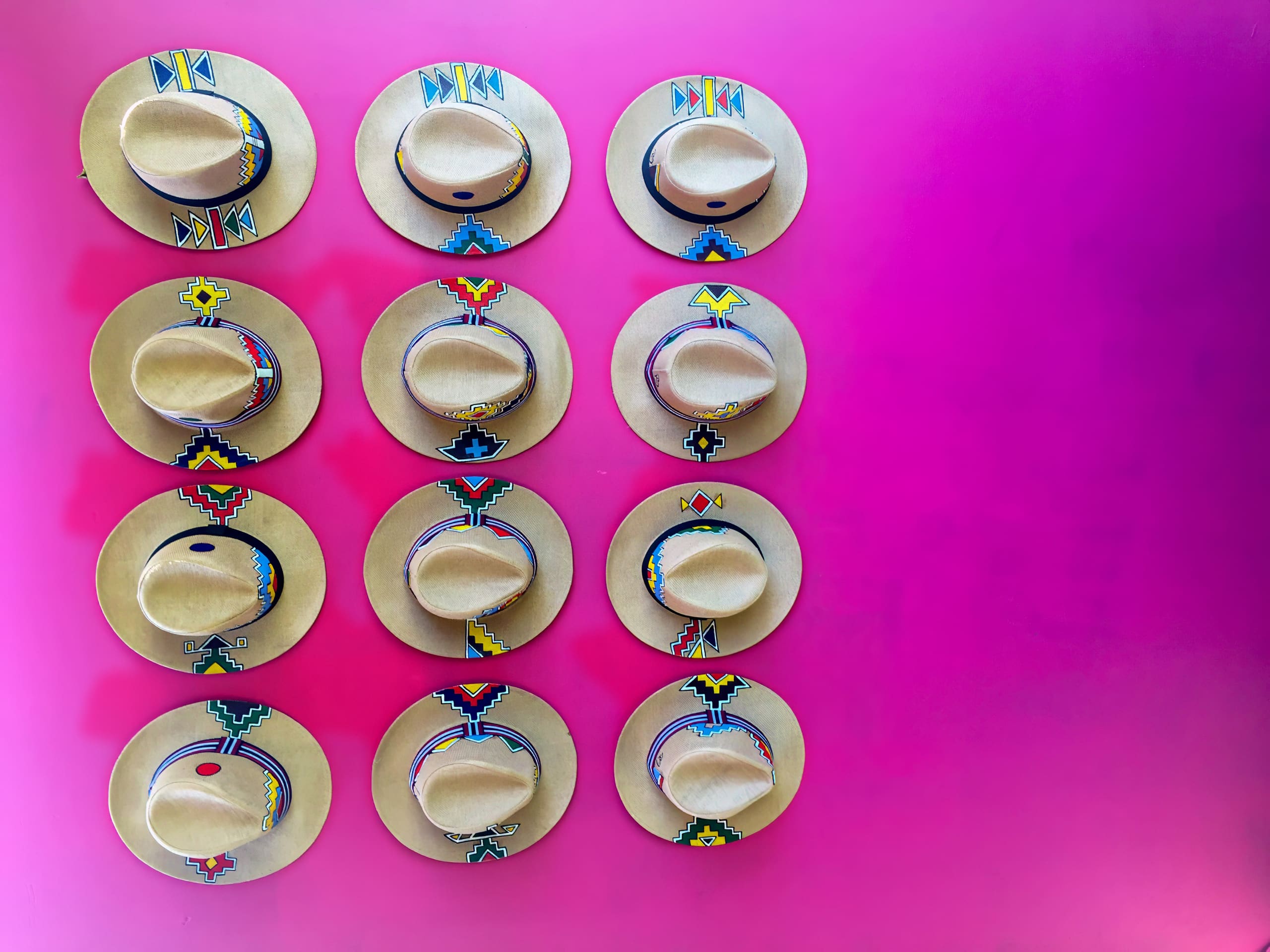Eleven beige hats decorated with colourful, intricate patterns are arranged in a grid on a bright pink wall, with each hat featuring unique beaded designs around the crown and brim. The vibrant pink background contrasts with the neutral colour of the hats.