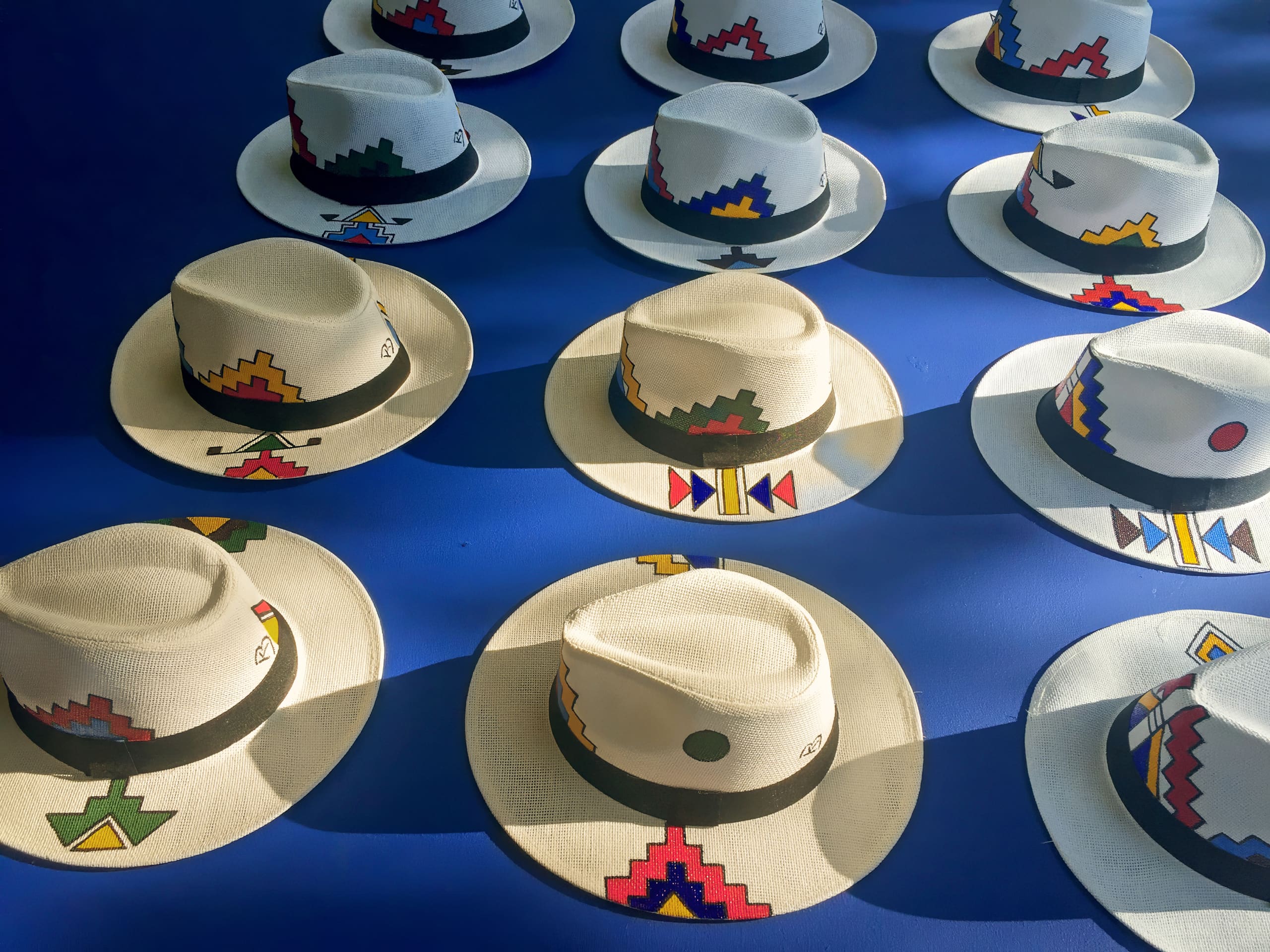 Fourteen white straw hats with colourful, geometric designs and black bands are arranged in rows on a bright blue surface. Sunlight casts shadows, highlighting each hat’s unique, vibrant pattern including reds, yellows, blues, and greens.
