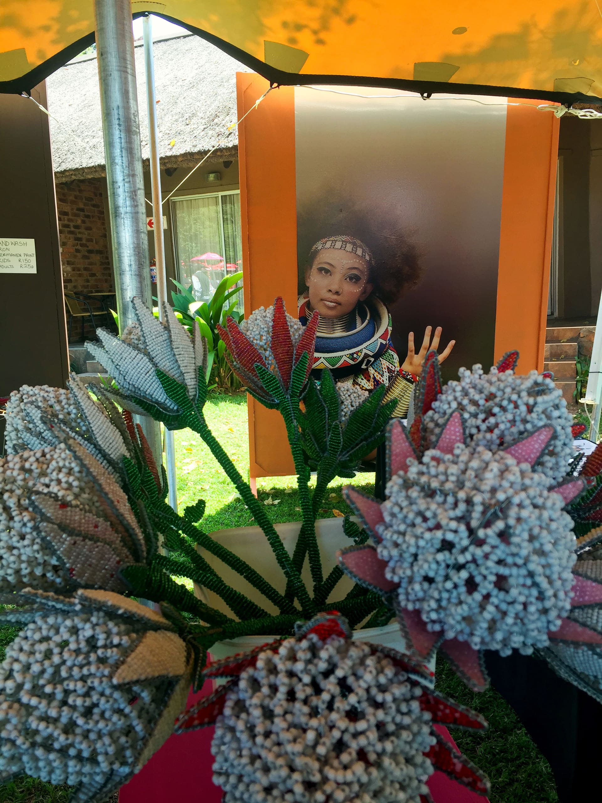 A bouquet of beaded protea flowers is displayed in the foreground. In the background, there is a vibrant portrait of a woman in traditional attire with a beaded necklace, raising her hands. The scene is set outdoors under a yellow canopy.