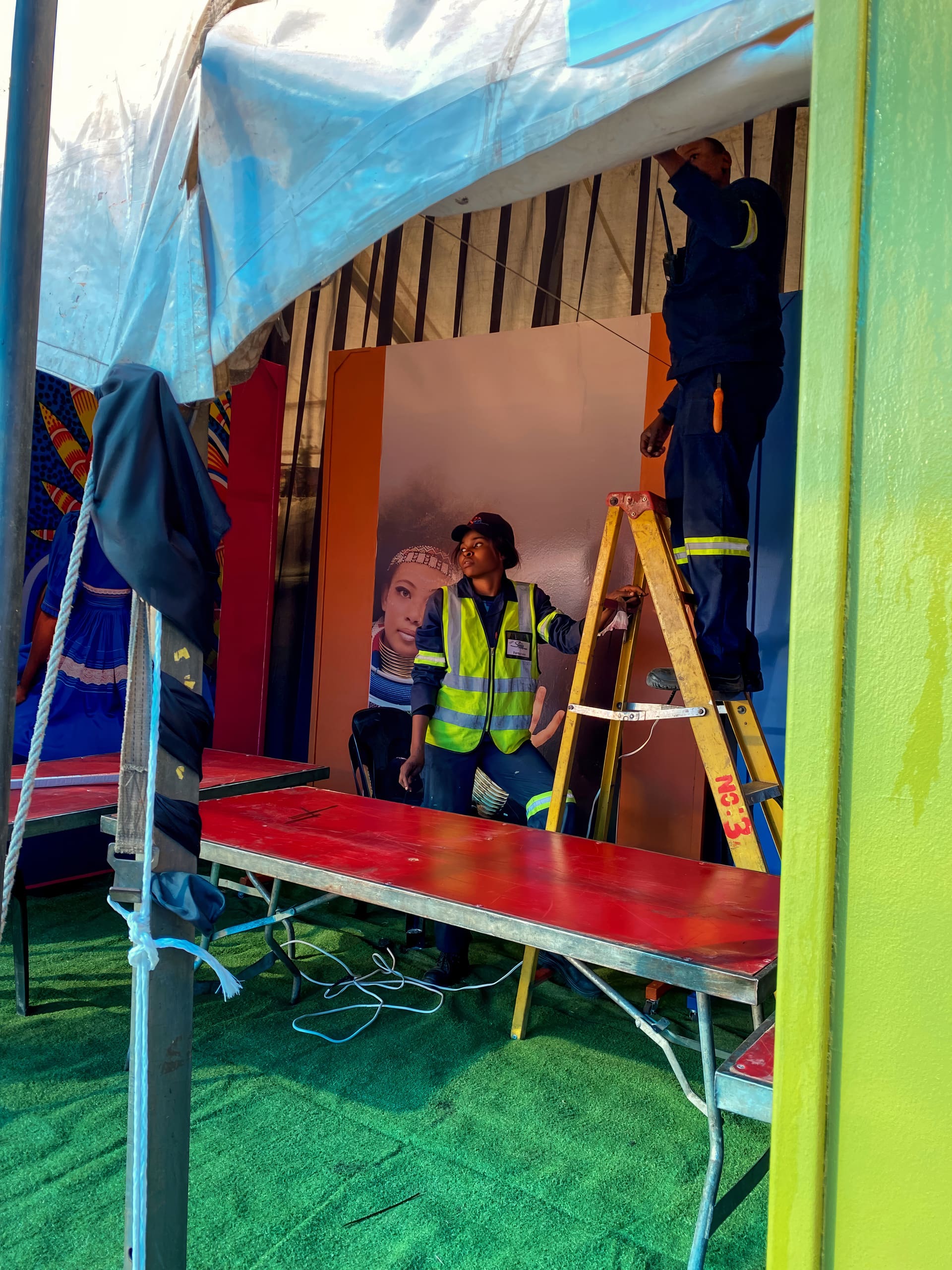Two workers in reflective vests and uniforms set up an indoor event stand. One stands on a yellow ladder adjusting the tent roof, while the other looks up, holding the ladder. Bright banners and portraits are visible behind them, with green artificial grass flooring.