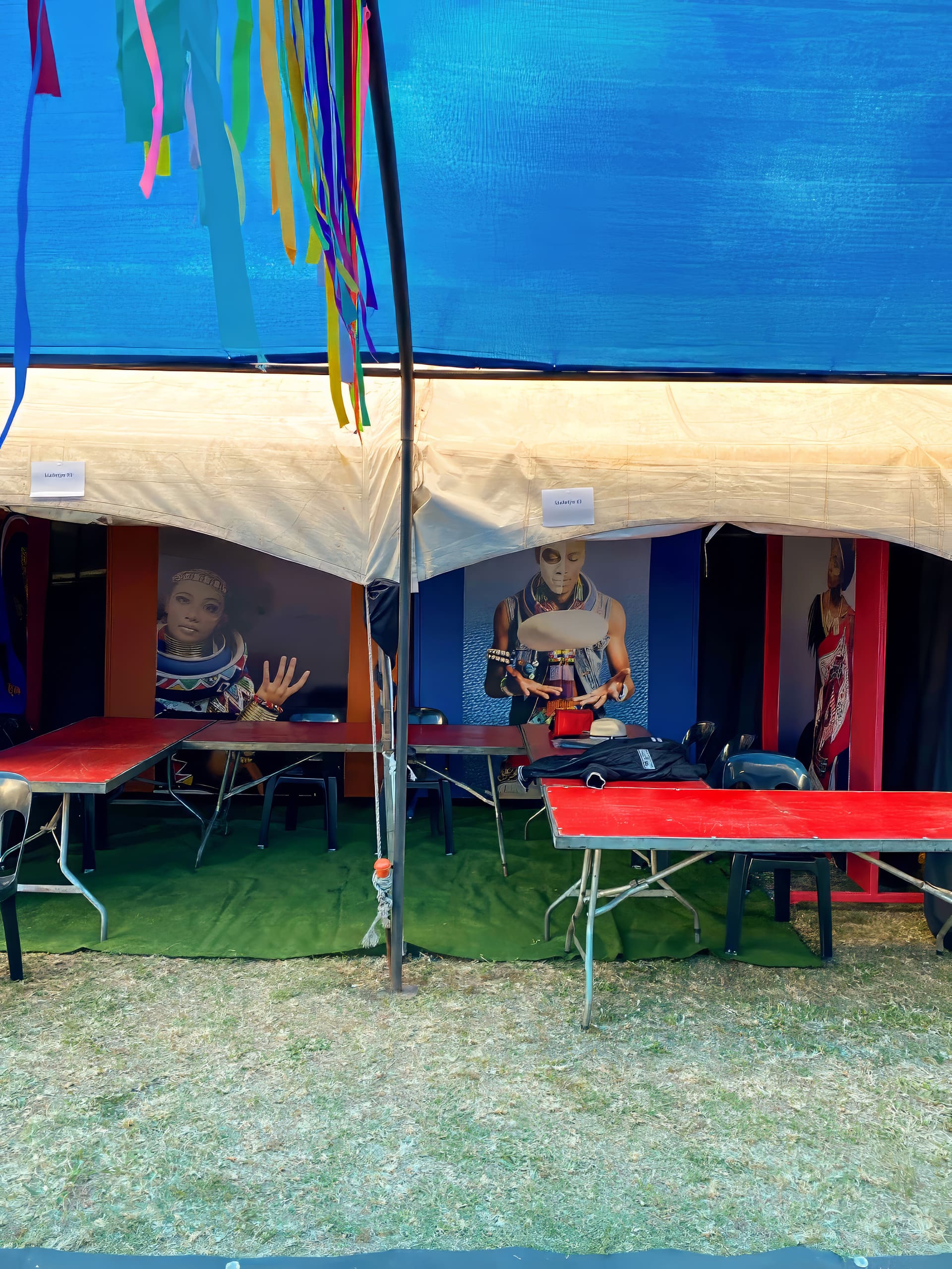 A tent with a beige roof and blue sides contains red tables and black chairs on green artificial grass. Colourful ribbons hang from a pole. Large posters of people in traditional attire decorate the tent’s back wall. The scene appears festive and lively.