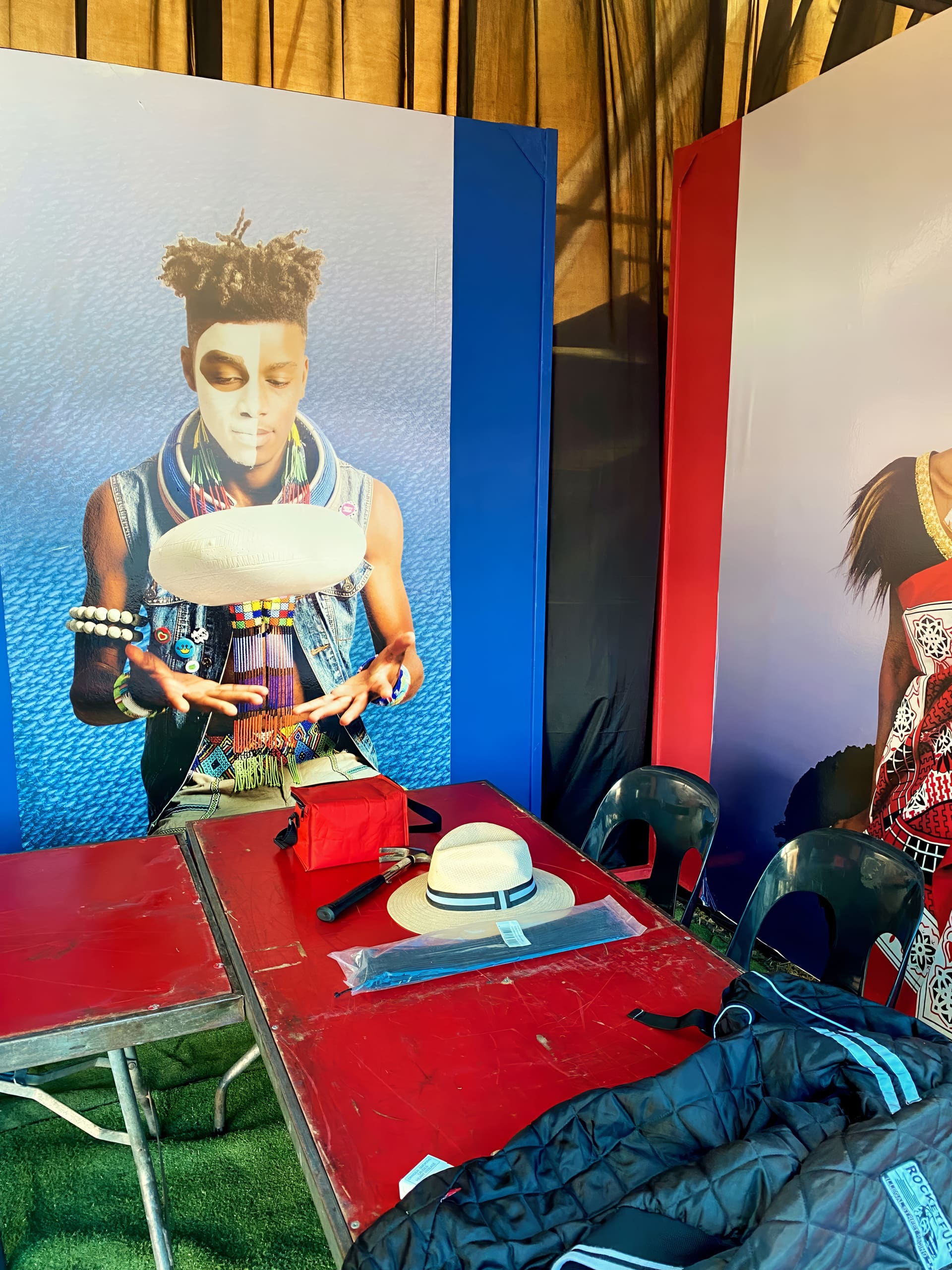 A colourful stall with a mural of a person in face paint, beads, and a denim waistcoat spinning a hat. In front, two red tables hold a beige hat, a red pouch, and a dark jacket; black chairs are around the tables, and part of another mural is visible on the right.