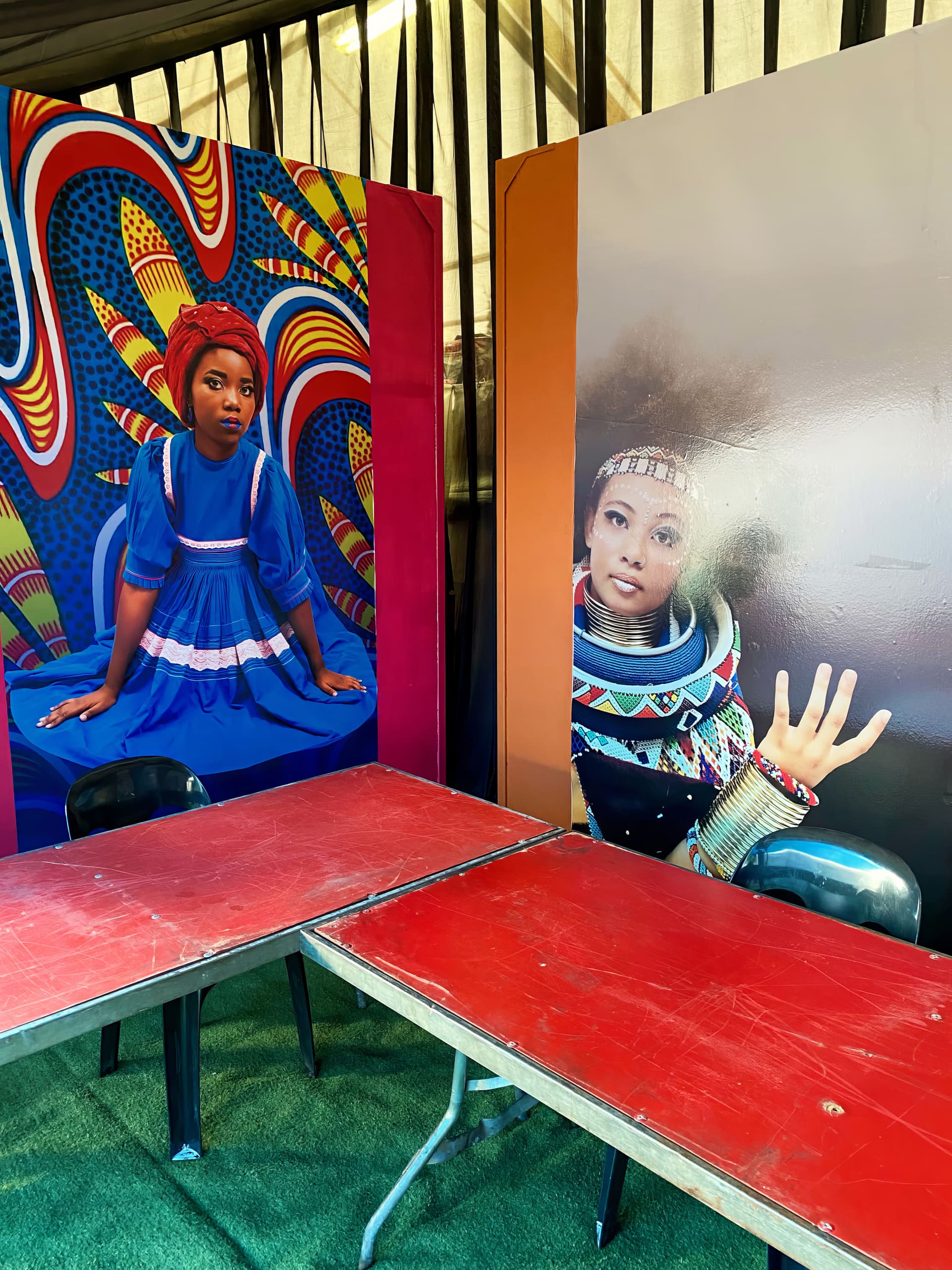 Two colourful portraits of Black women are displayed behind red tables and black chairs. One woman wears a blue dress and patterned headwrap, seated before a vibrant abstract background. The other is in futuristic attire, raising her hand in a gesture.