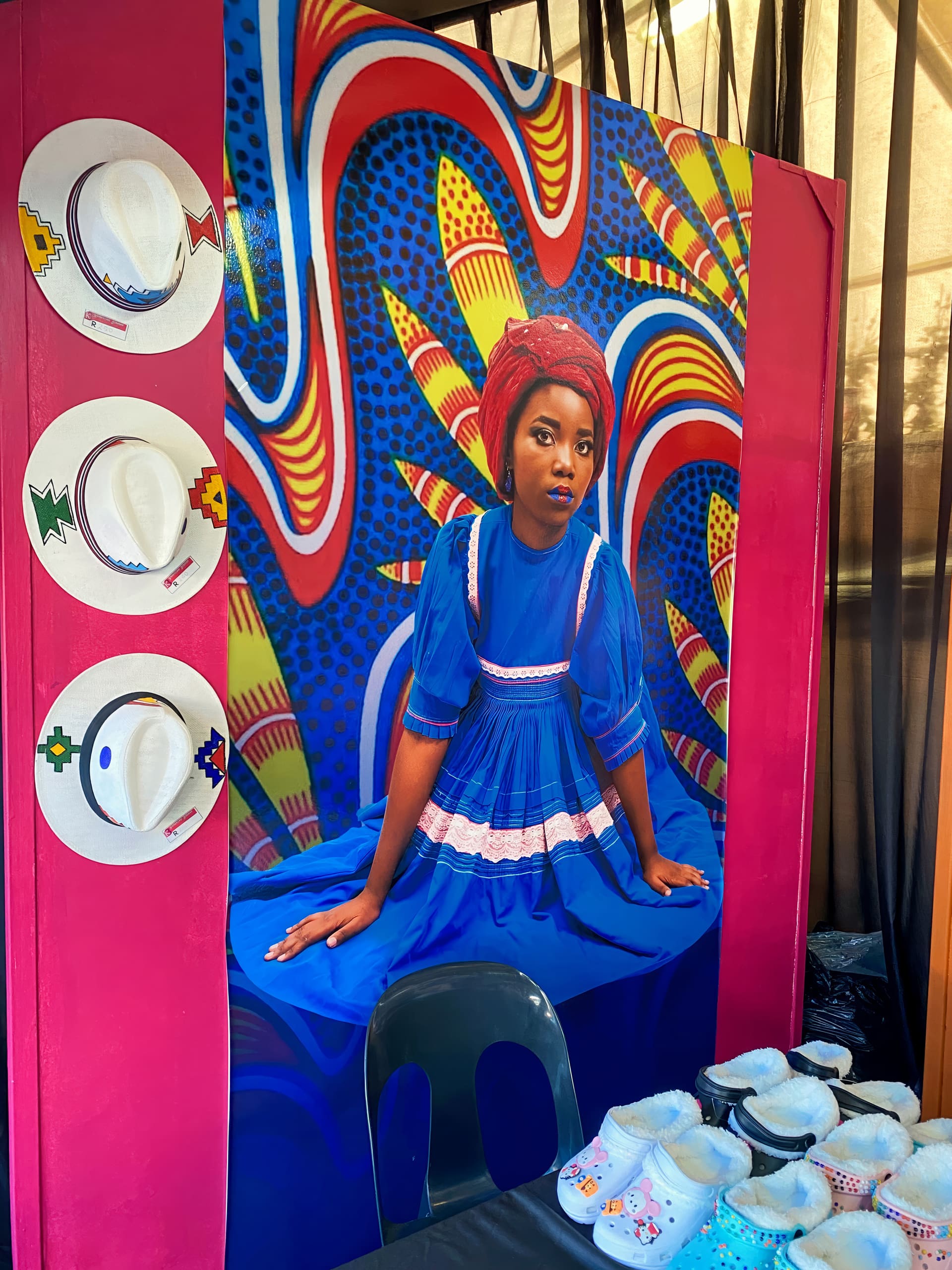 A vibrant display features a portrait of a woman in a blue dress and red headscarf, set against a colourful, abstract patterned backdrop. To the left, three decorated white hats hang on a pink board. Baby shoes are arranged at the bottom right.