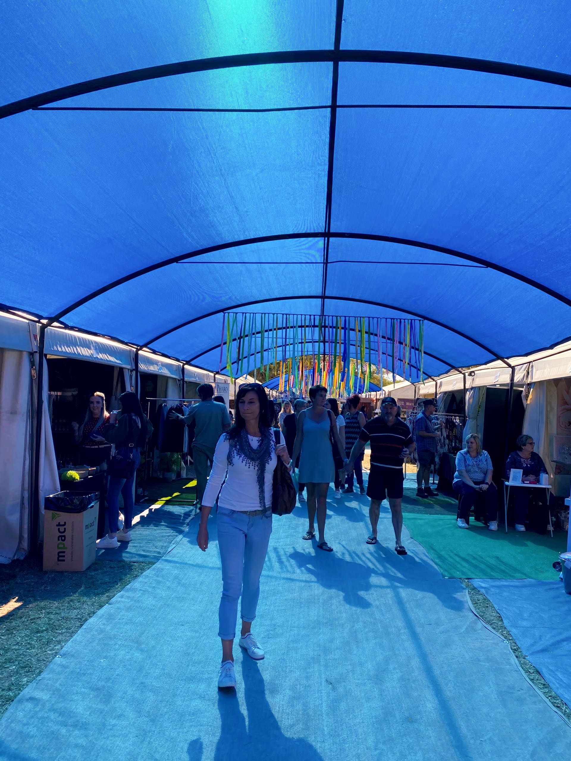 People walk and browse under a large blue canopy at an outdoor market or fair. Stalls line both sides, selling goods, as shoppers mingle and vendors sit. Colourful ribbons hang at the far end, adding a festive atmosphere. Sunlight filters softly through the canopy.