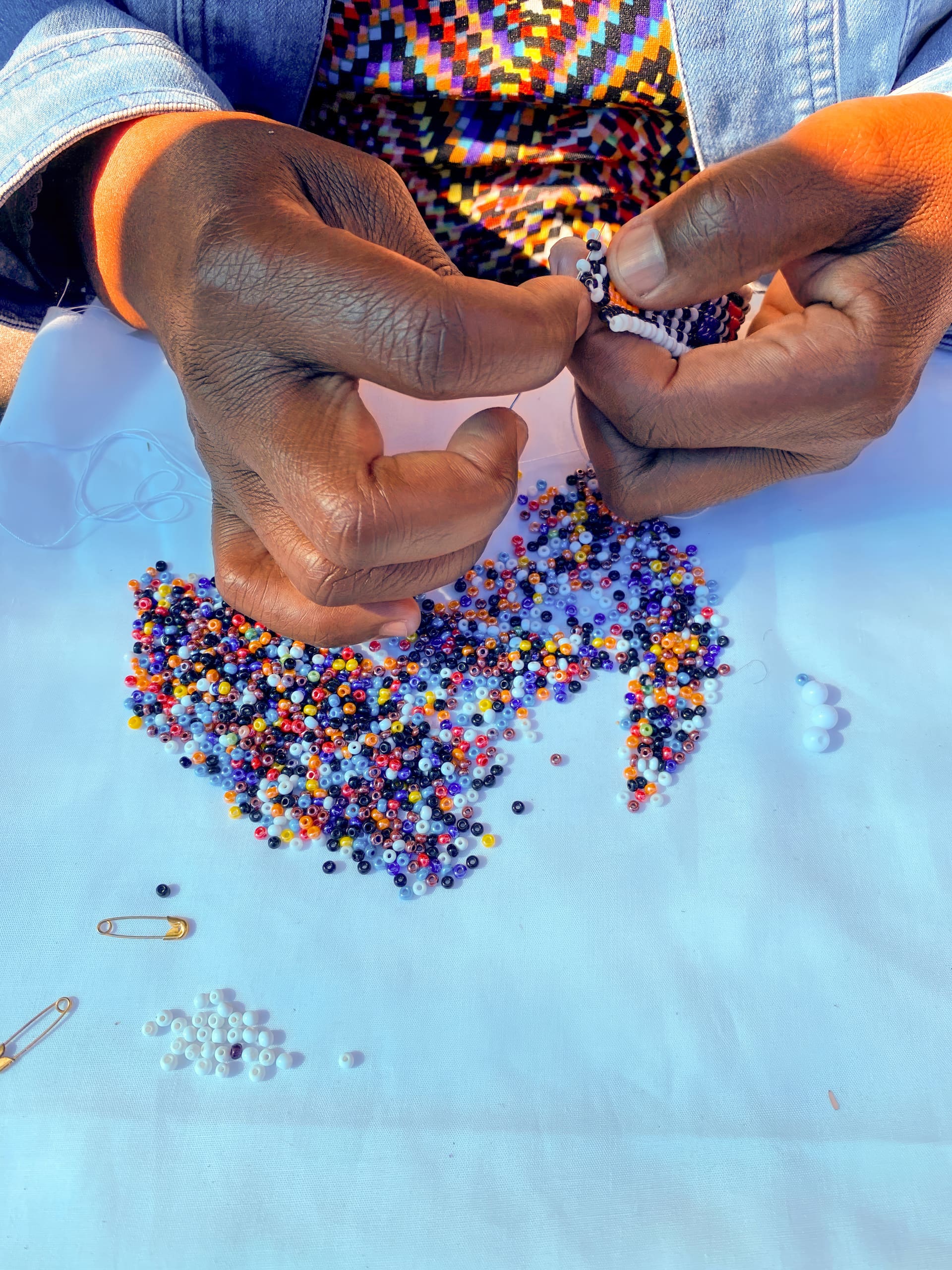A person’s hands thread colourful beads onto a string over a white surface. Mixed beads in various colours—purple, yellow, red, white, blue, and black—are scattered across the table. The person wears a patterned top and a denim jacket.