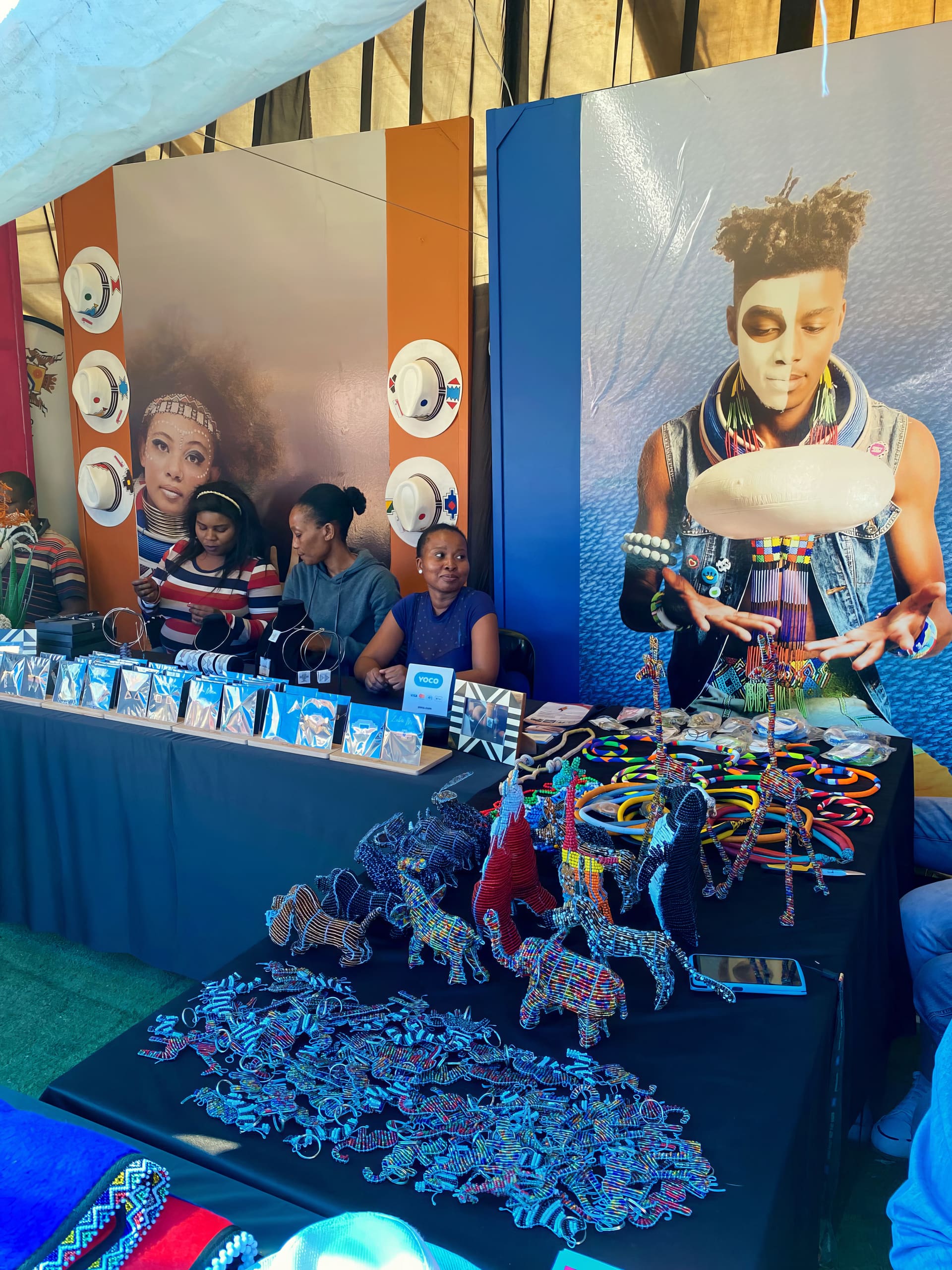 A stall at a market features three women behind tables displaying colourful beaded jewellery, animal figurines, and crafts. Behind them are large posters showing people in traditional attire and jewellery, with vibrant patterns and deep blue and orange backgrounds.
