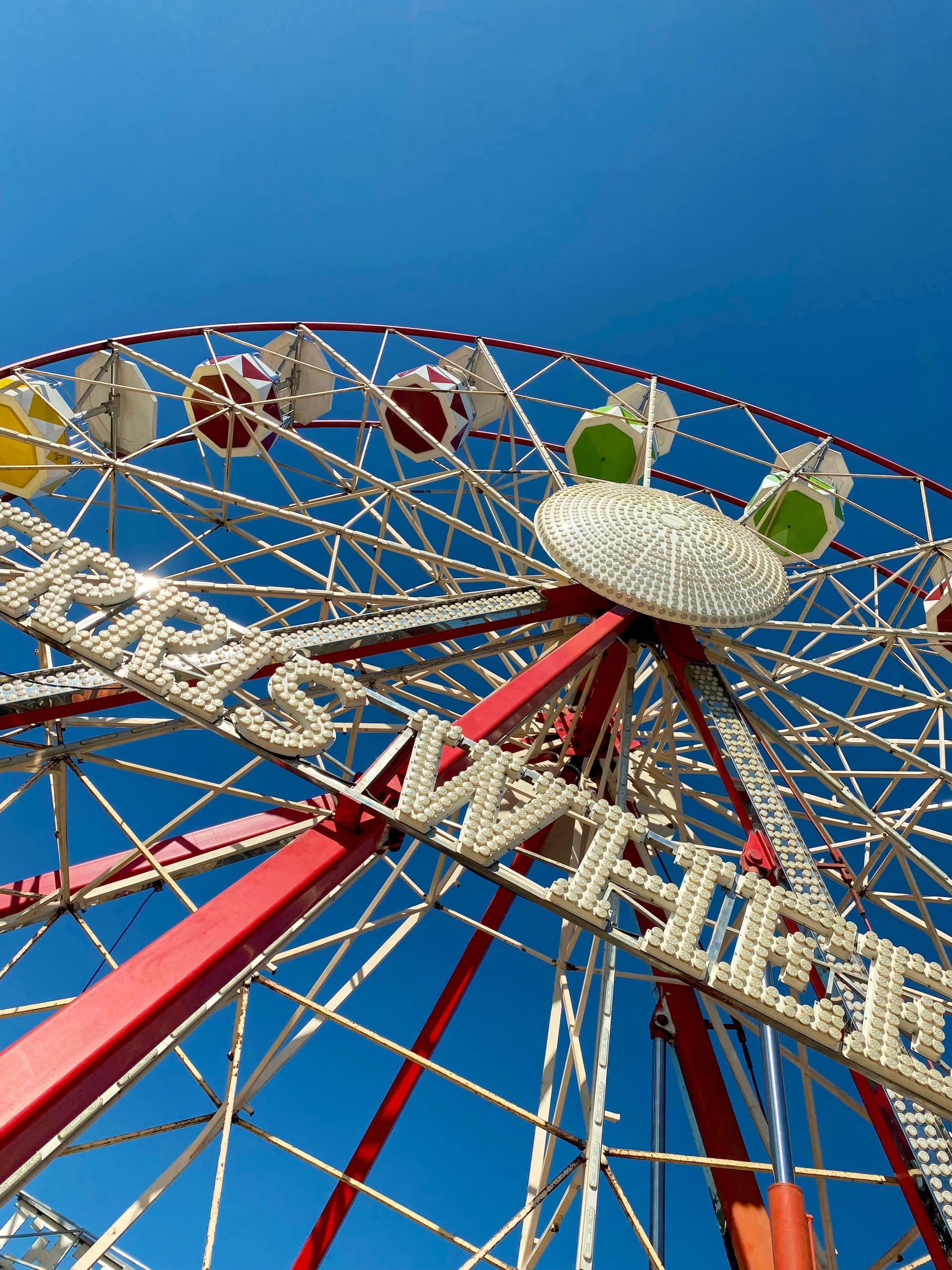 A close-up upward view of a Ferris wheel against a clear blue sky, showing red and white spokes, colourful gondolas, and part of a sign with large white letters, partially reading “WHEEL.”.