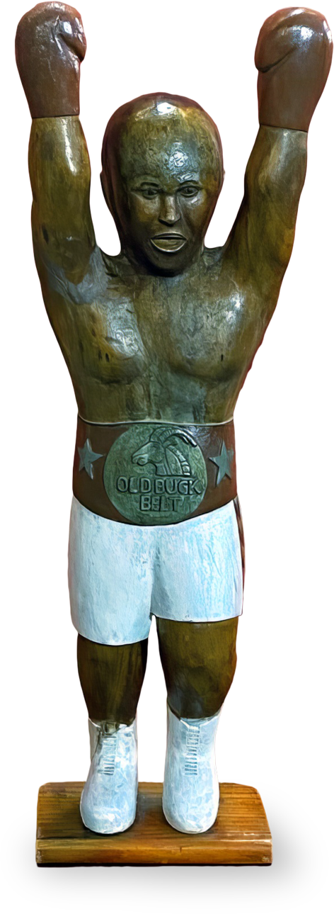 A wooden sculpture of a boxer with arms raised triumphantly. The figure wears white boxing shorts, white shoes, and a large silver belt reading “OLD DOG BELT.” The sculpture is mounted on a rectangular wooden base.