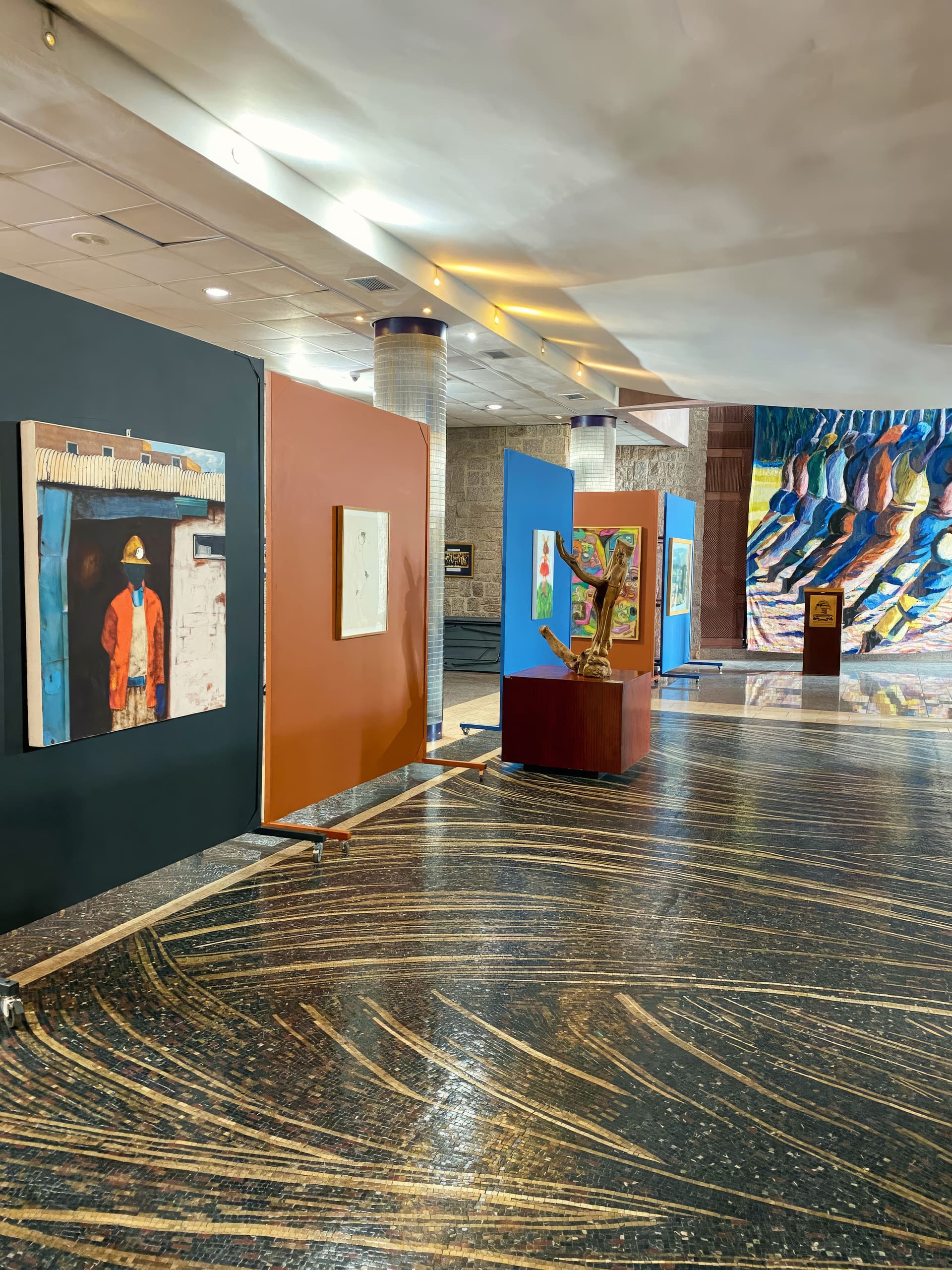 Indoor art exhibition with several colourful paintings displayed on freestanding panels. The floor has curving, striped patterns. In the background, a large wall mural with abstract blue shapes is visible. Lighting is warm and soft throughout the space.