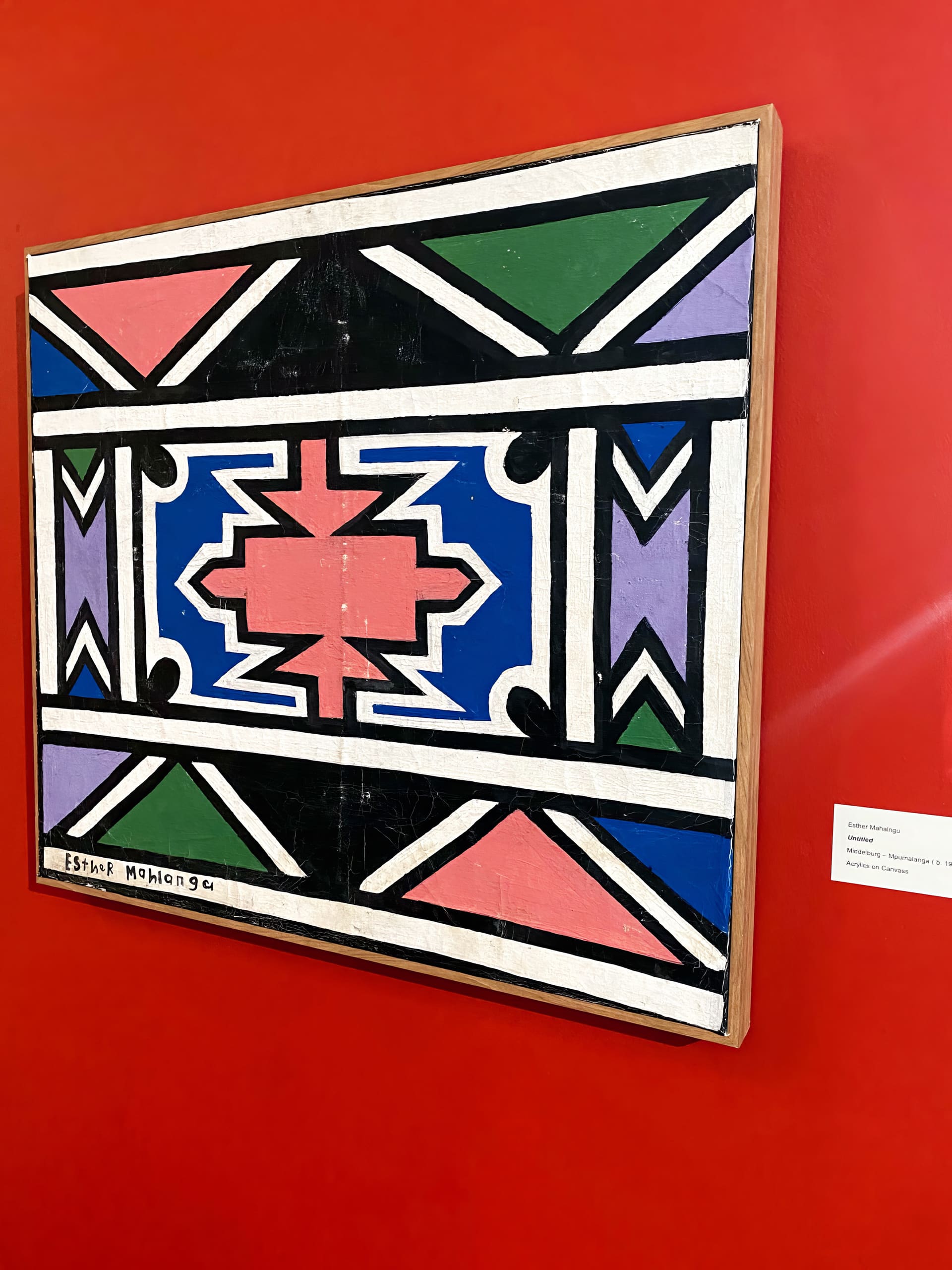 A geometric painting in bold colours—blue, pink, green, black, and white—featuring symmetrical patterns and shapes, hangs on a vibrant red wall. The artist’s signature, Esther Mahlangu, is visible in the lower left corner. A small label is mounted nearby.