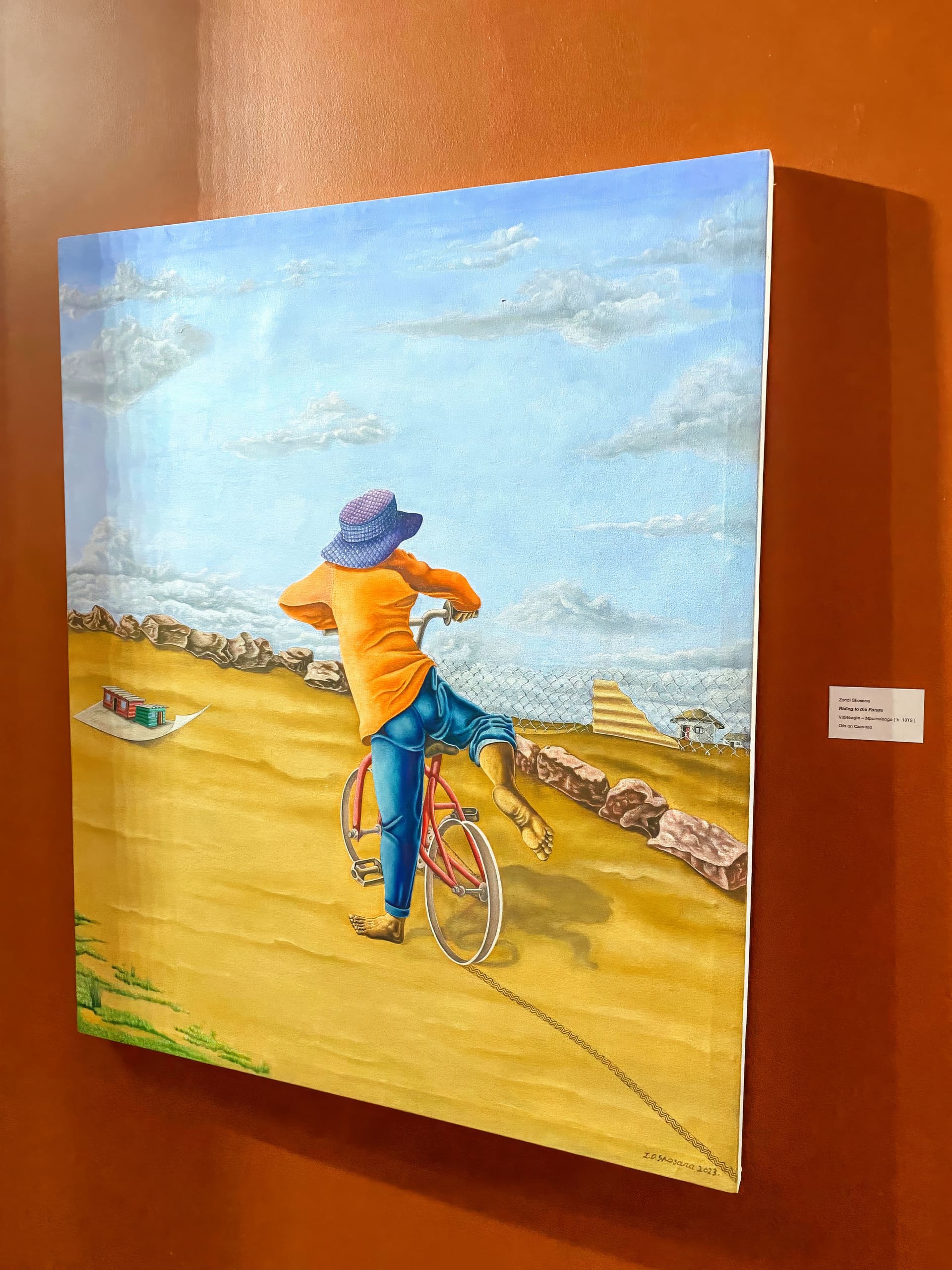 A colourful painting on a wall shows a person in an orange shirt, blue trousers, and a blue hat riding a pink bicycle on a sandy path, kicking one leg up playfully. The sky is blue with clouds and a fence and low wall appear in the background.