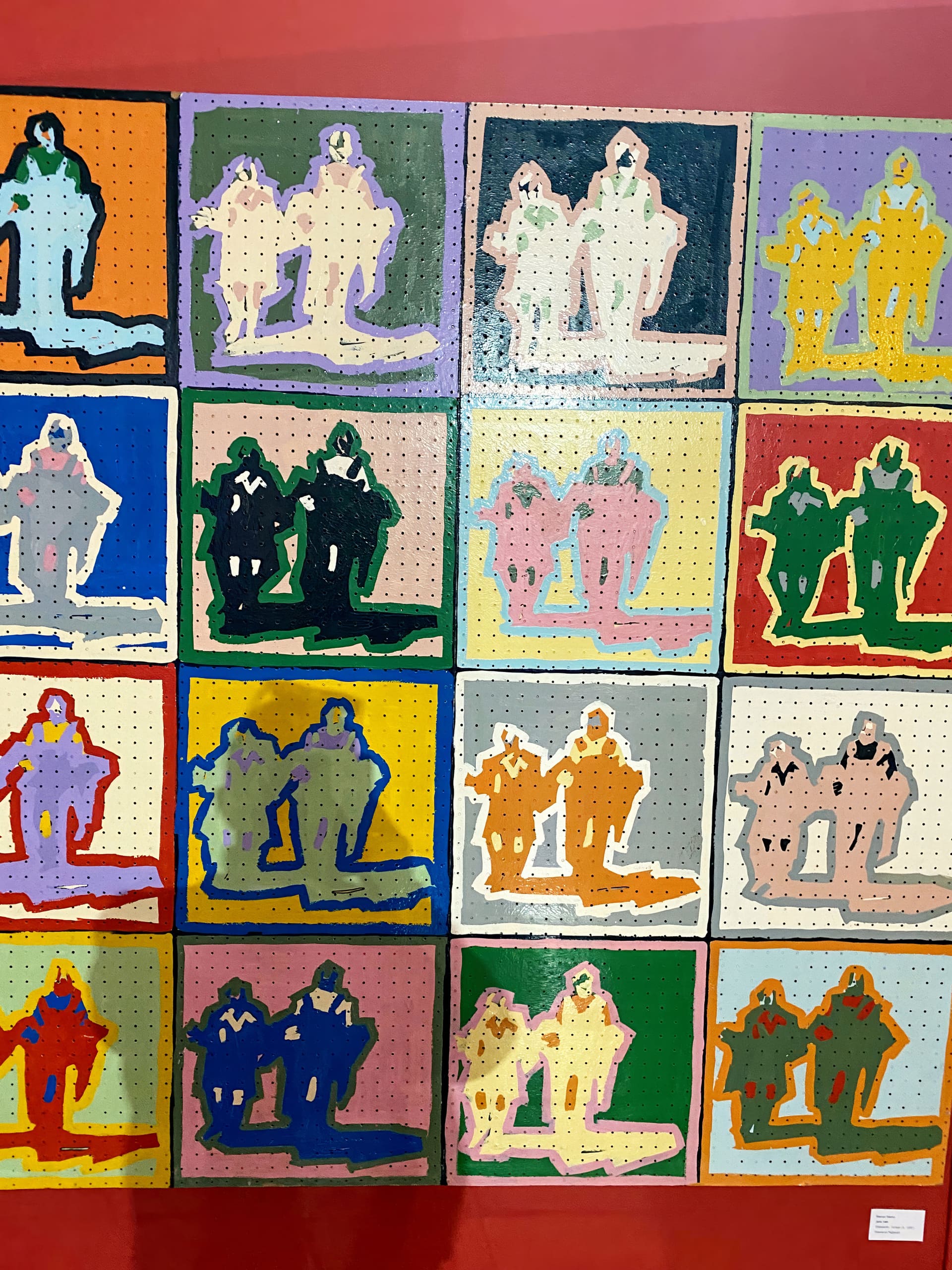 A grid of colourful pop-art style paintings, each square showing two abstract, human-like figures kneeling side by side. The background and figures vary in bold colours and outlines, arranged against a red wall. The patterns resemble Andy Warhol’s repetitive style.