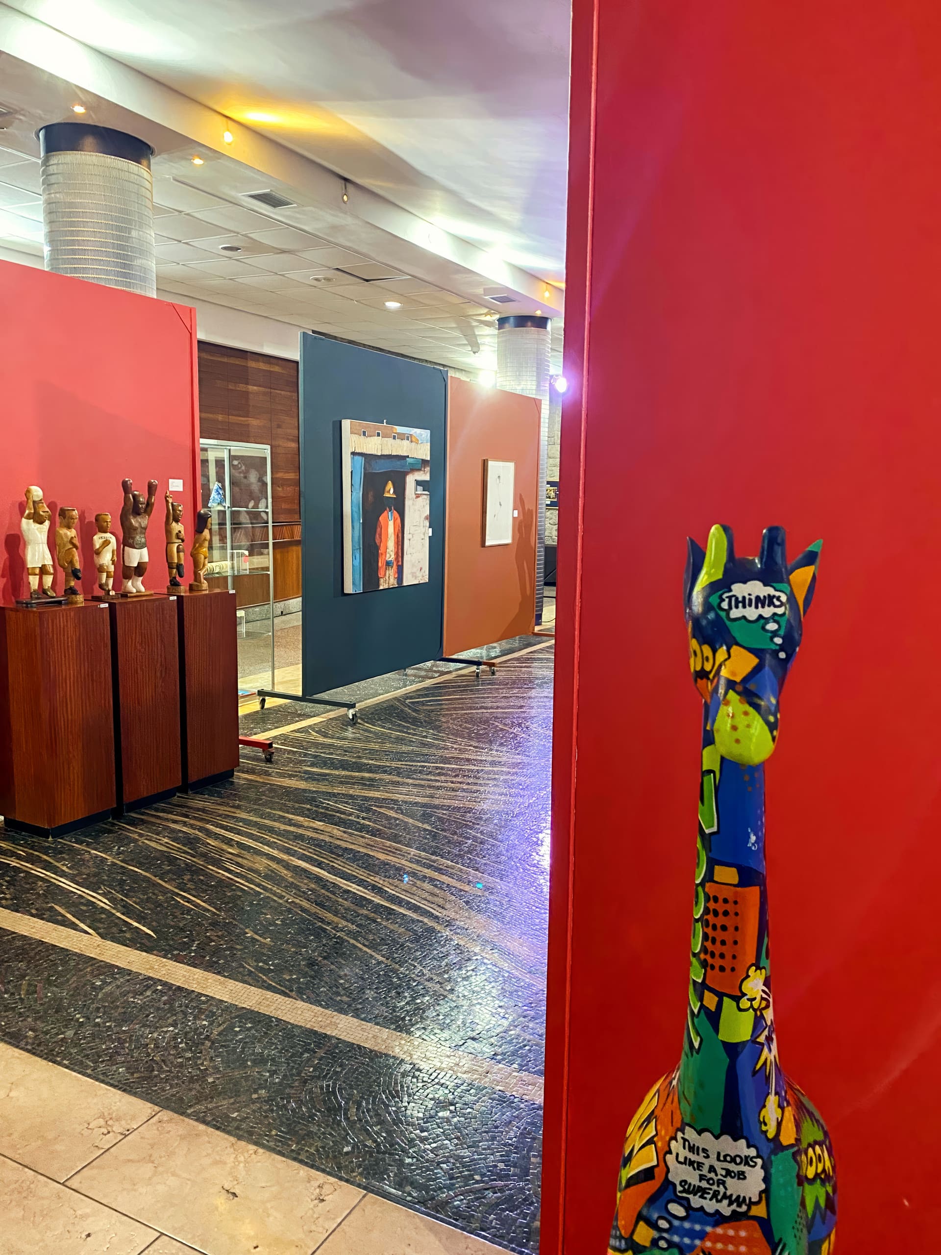 A colourful, comic-themed giraffe sculpture stands near a red wall at an art gallery. In the background, sculptures and framed artworks are displayed on stands and panels under warm lighting, with a glossy floor and modern decor visible.