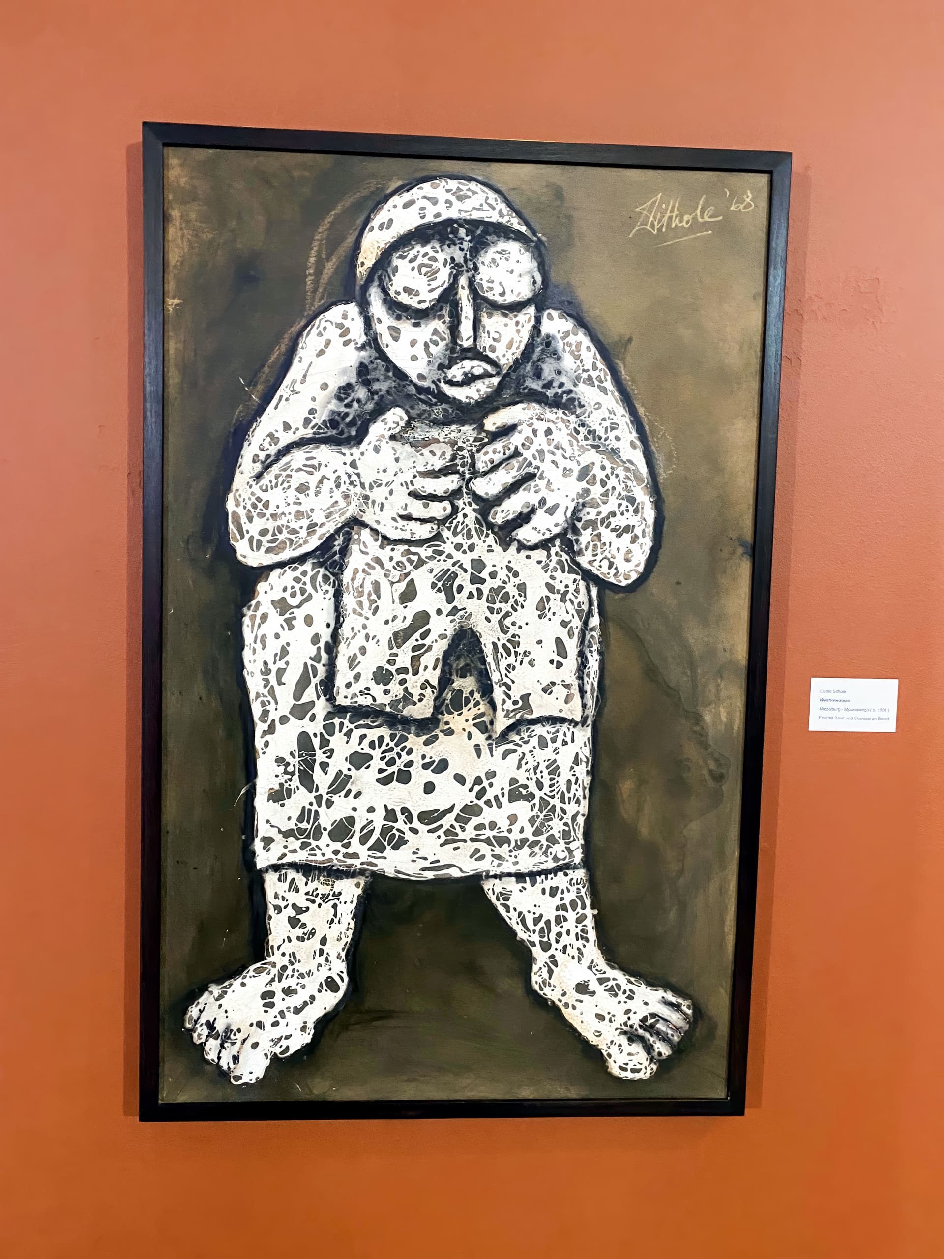 A painting shows a stylised, crouched figure with oversized feet and hands, textured with white and black patterns. The figure wears glasses and looks forward. The background is a muted brown, and the artwork hangs on a burnt orange wall.