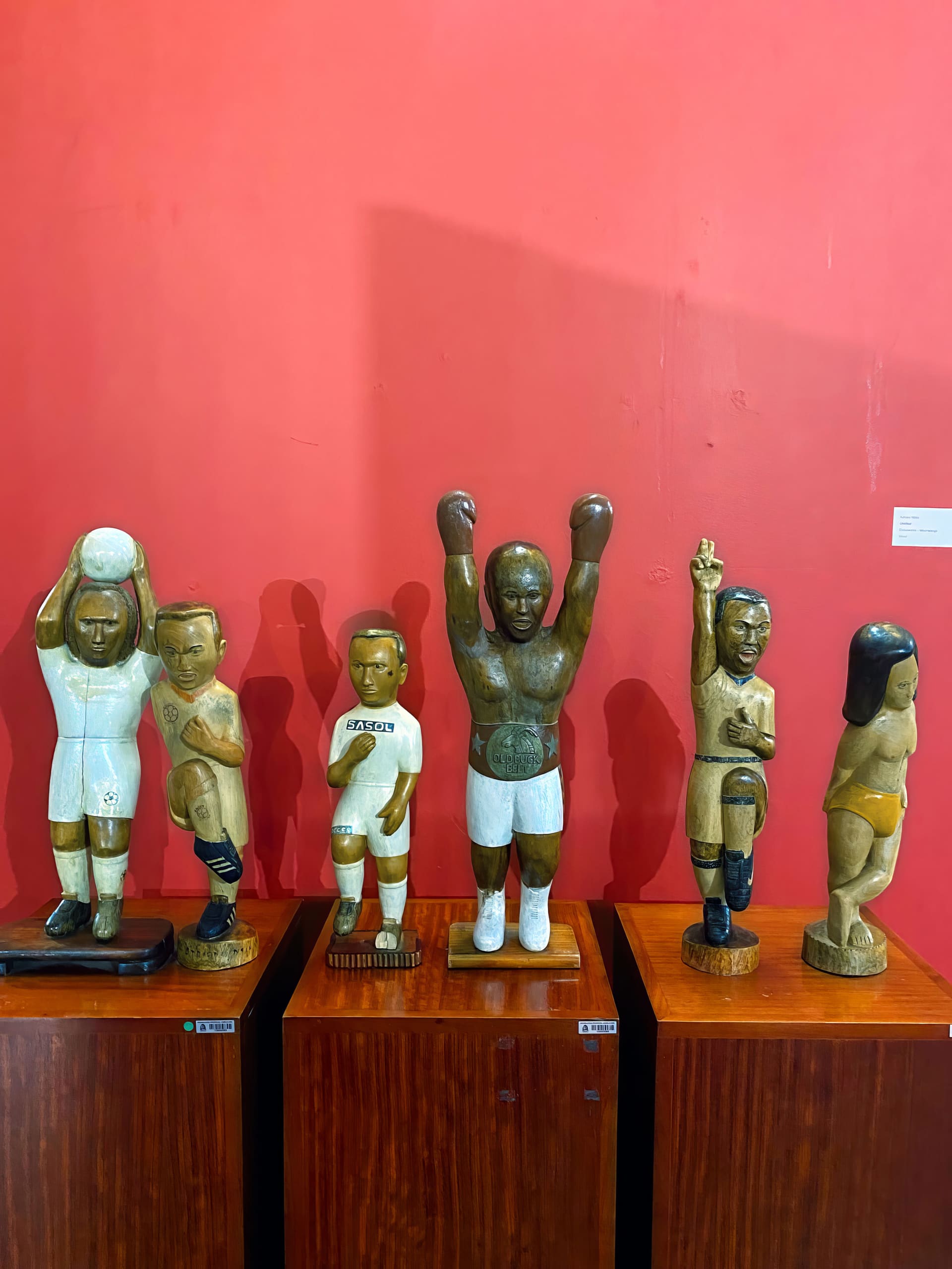 Five carved wooden statues of athletes on brown plinths stand against a solid red wall. The figures resemble footballers and a boxer with his arms raised, each with simplified human features and wearing sports kits.