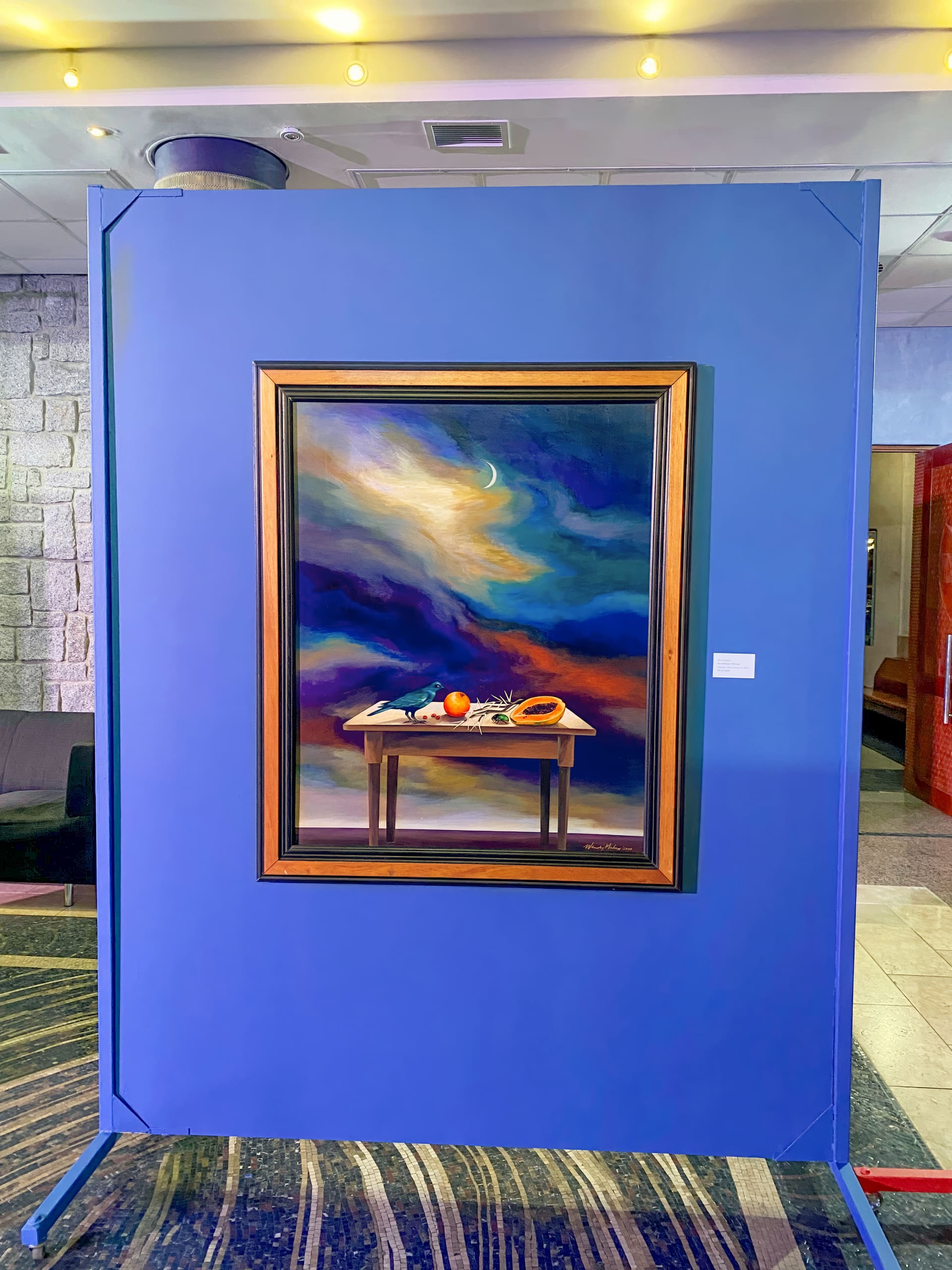 A framed painting on a blue display panel depicts a table with fruit and objects under a dramatic, swirling night sky. The scene is inside a well-lit modern space with stone walls, striped carpet, and a red partition visible in the background.