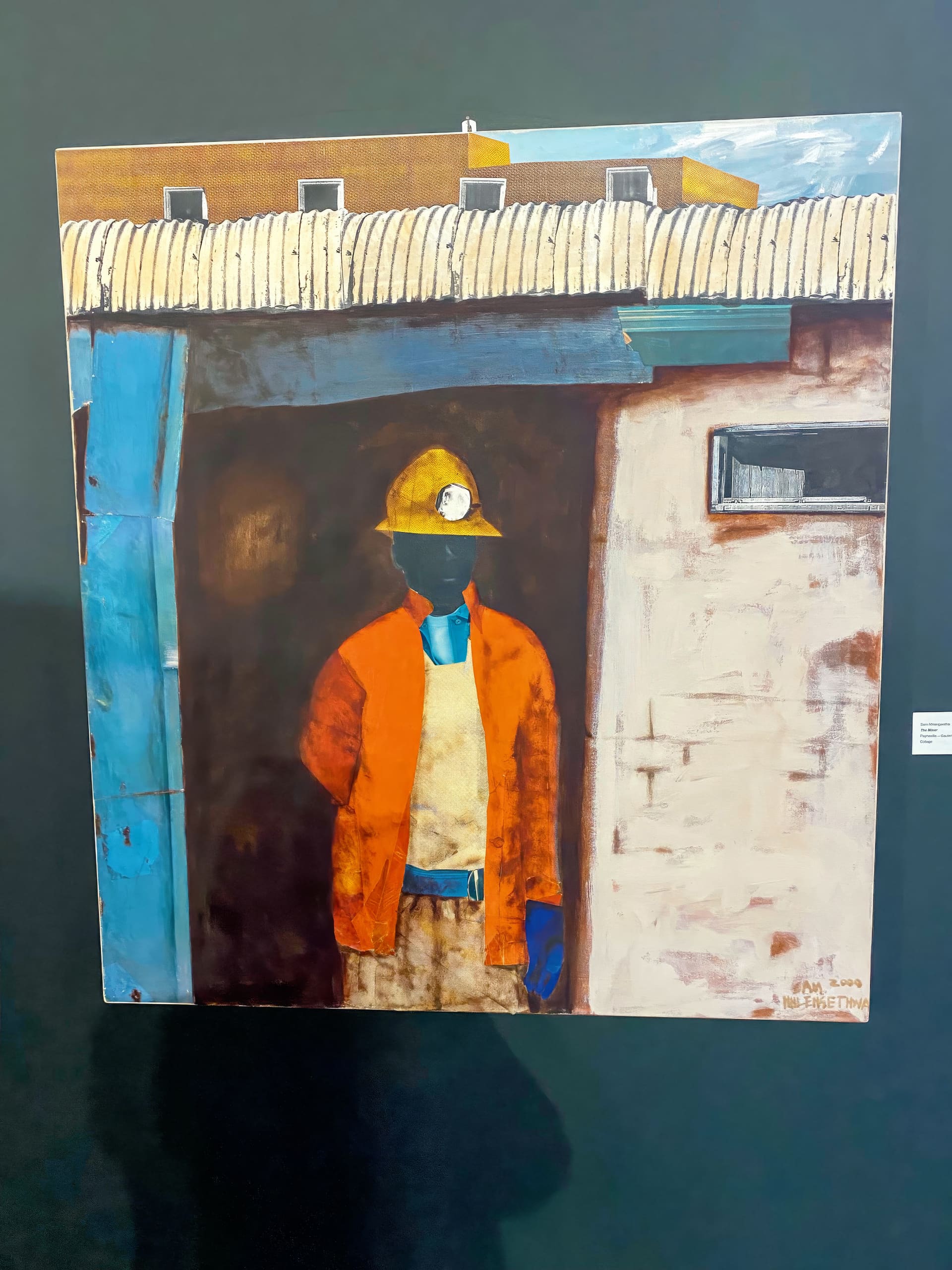 A painting of a figure in an orange jacket, yellow hard hat with headtorch, and blue gloves stands partly in shadow by a building with blue and white walls under a curved metal roof. The figure’s face is obscured by shadow. The background shows a yellow structure.