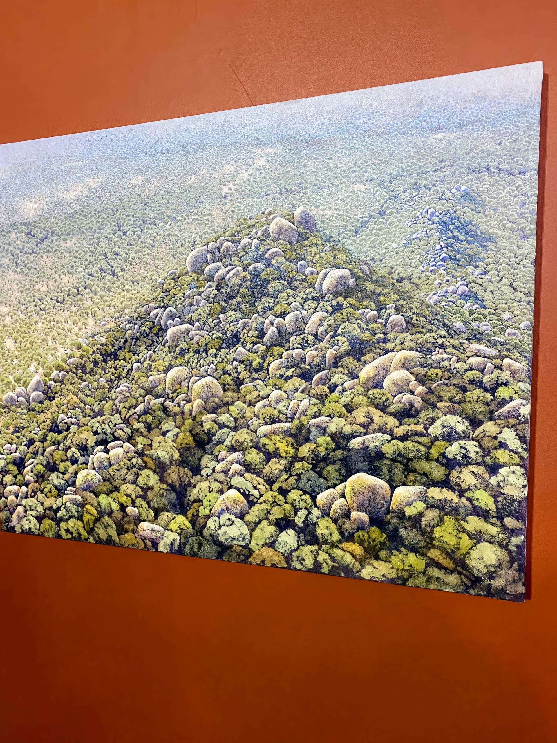 A painting of a lush, green forested landscape with rounded hills covered in dense trees and large boulders, mounted on a burnt orange wall. The upper right corner of the artwork is not visible.