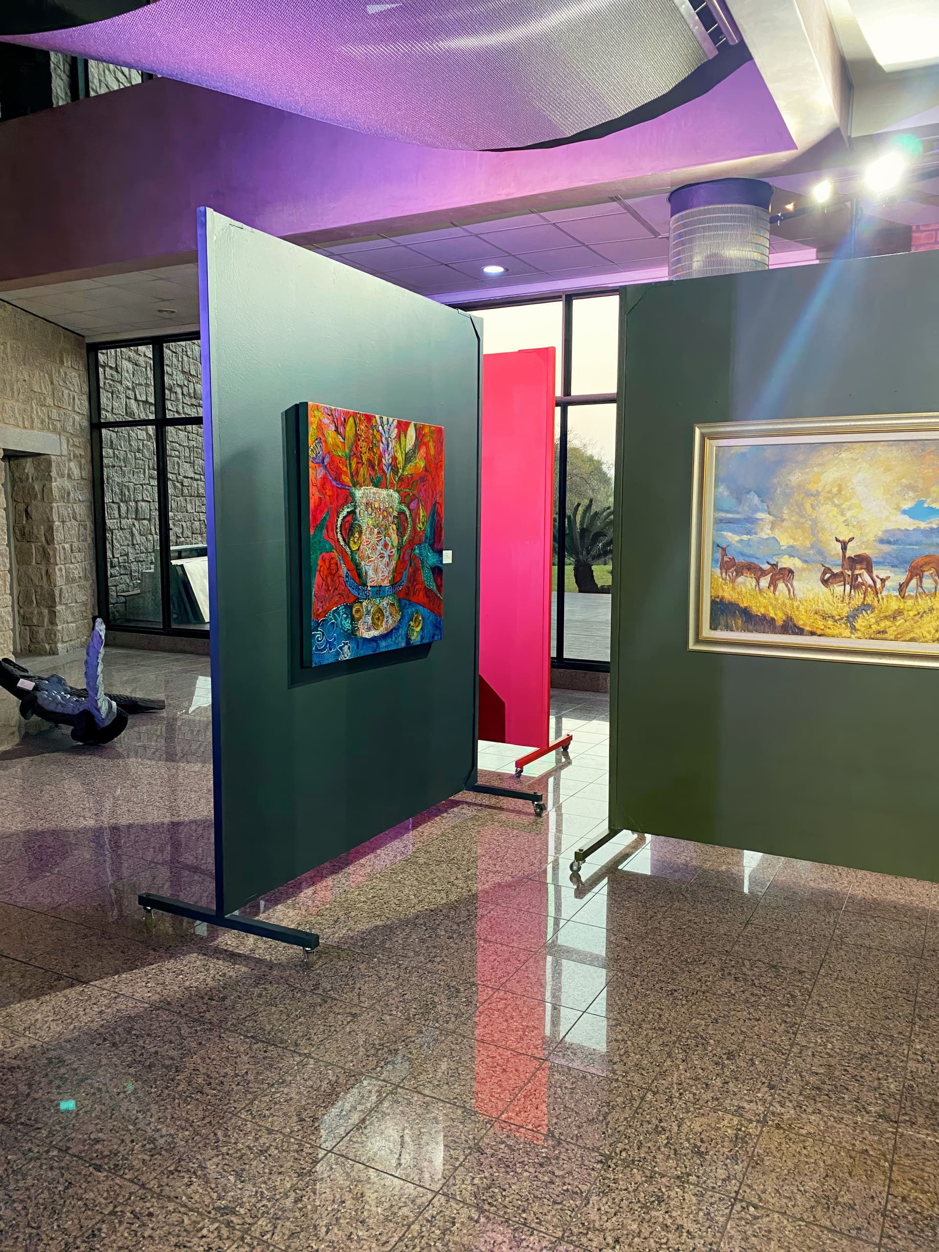 An art gallery with glossy tiled floors displays colourful paintings on wheeled partitions. One painting shows a vibrant floral arrangement in a vase; another depicts antelopes on a savanna. Sunlight streams in from large windows. A crocodile sculpture sits in the background.