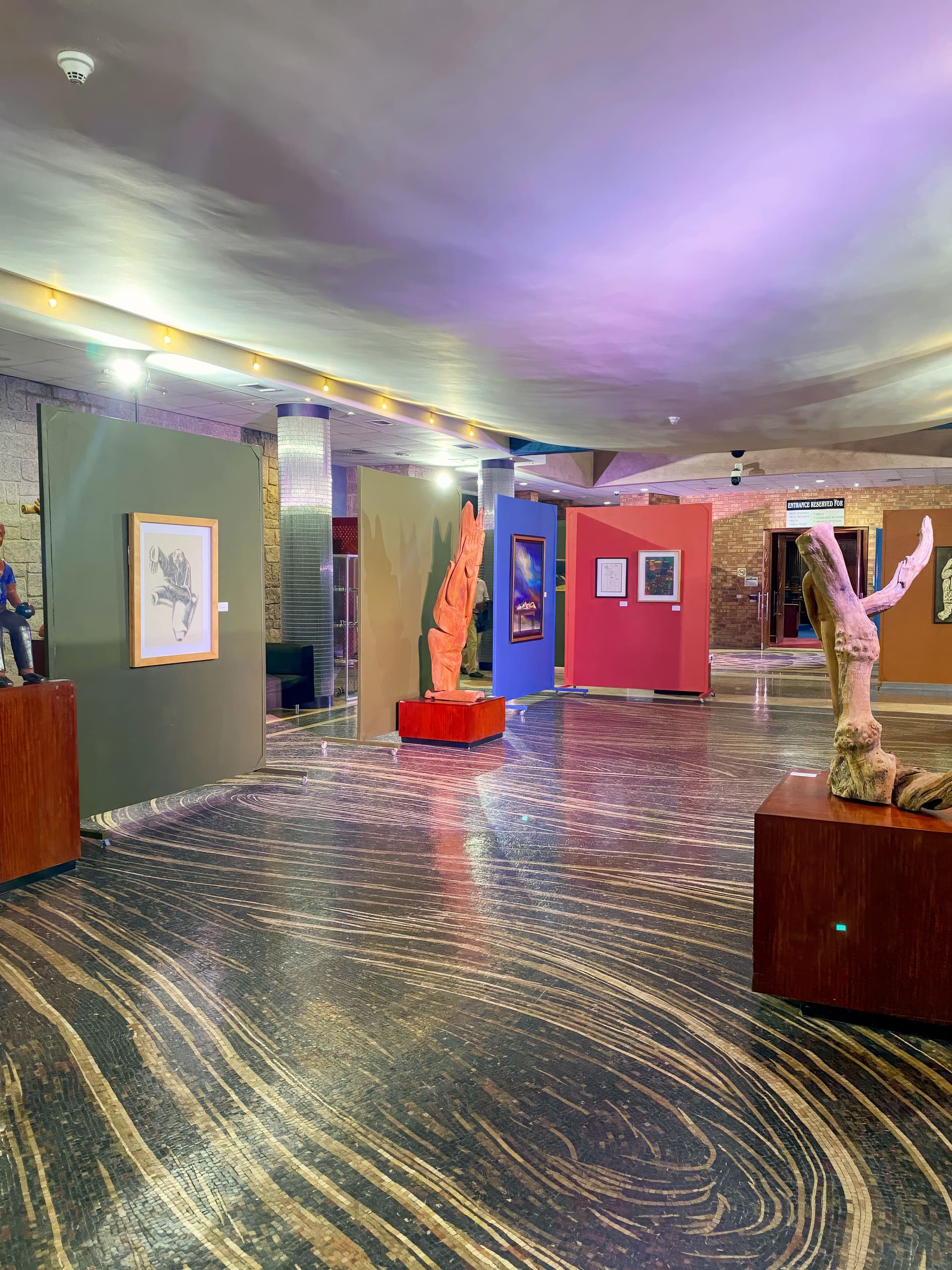 A museum gallery with wavy-patterned floors, colourful display walls with framed art, and sculpted wooden pieces on plinths. Soft overhead lighting and a mix of wall colours create a warm, modern atmosphere. The room appears empty of visitors.