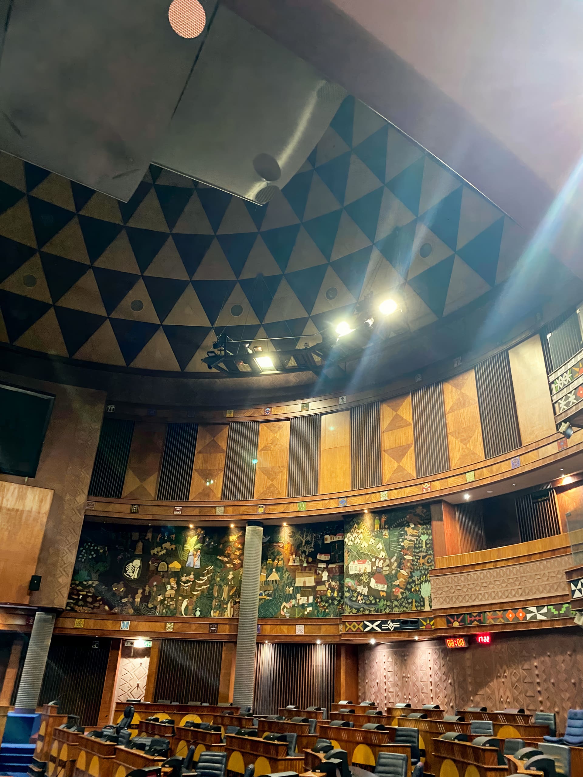 A curved legislative chamber with wood-panelled walls, geometric patterns, and a painted mural. Rows of wooden desks with chairs face forward. The ceiling features dark triangles and bright lights. The atmosphere is formal and grand.