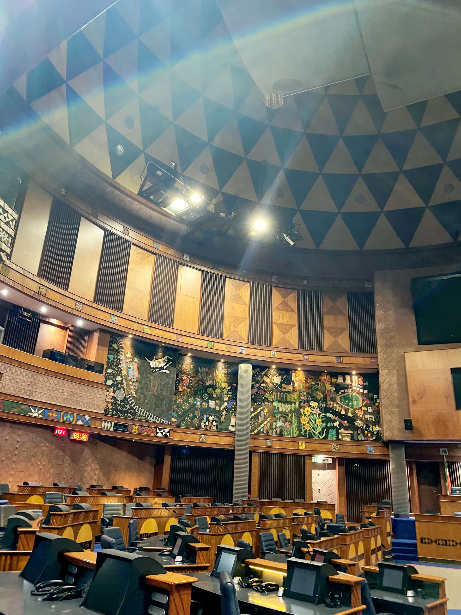 A spacious, circular parliamentary chamber with wooden desks and chairs arranged in rows. The walls feature vibrant murals, African motifs, and geometric patterns. Soft lighting and a high, triangular-patterned dome ceiling create an official, grand atmosphere.