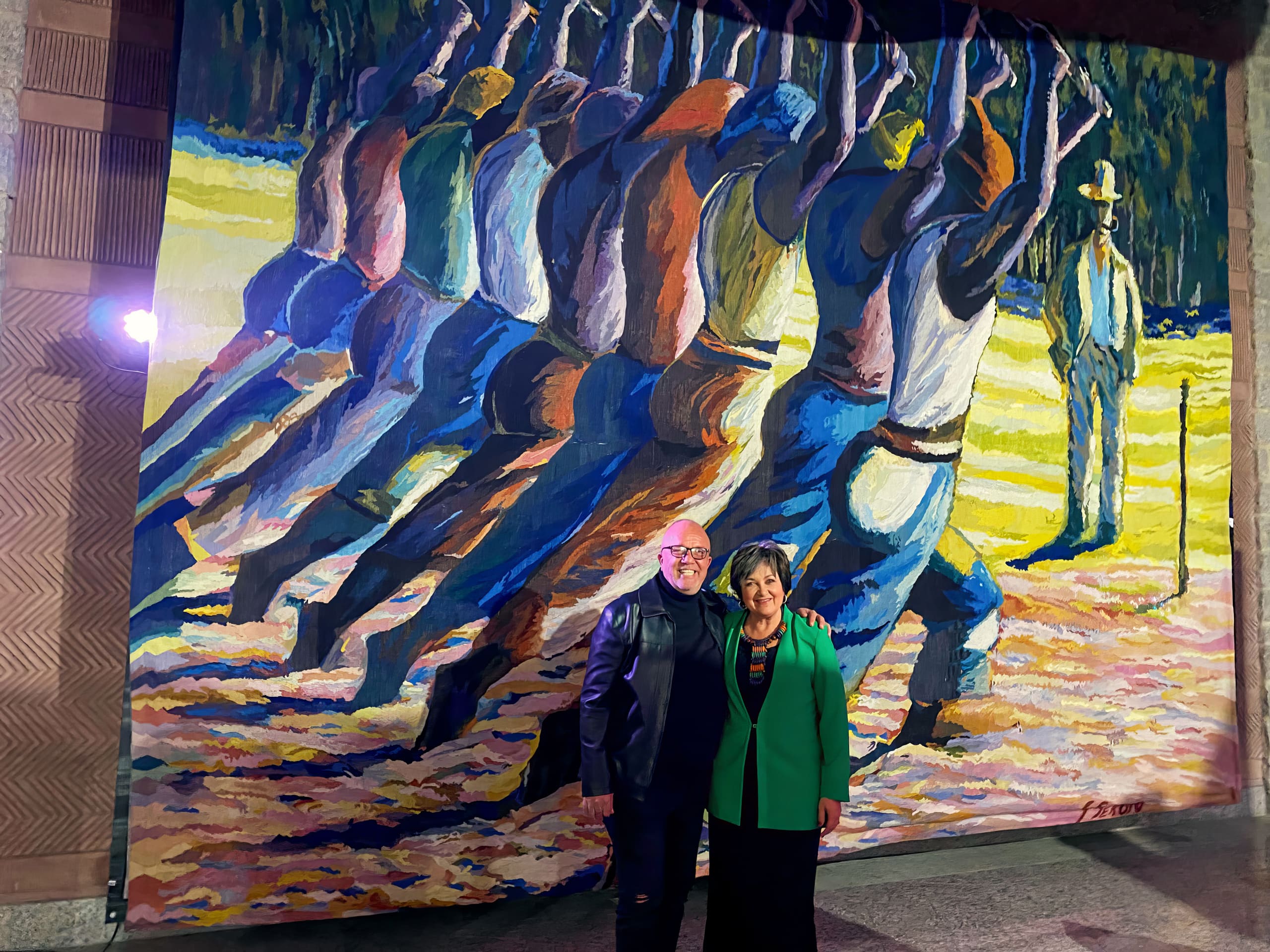 Two people stand smiling in front of a vibrant mural. The mural depicts a group of workers, arms raised, painted in bold colours. A single figure stands off to the right. The people in front are well-dressed and posed together under bright lighting.