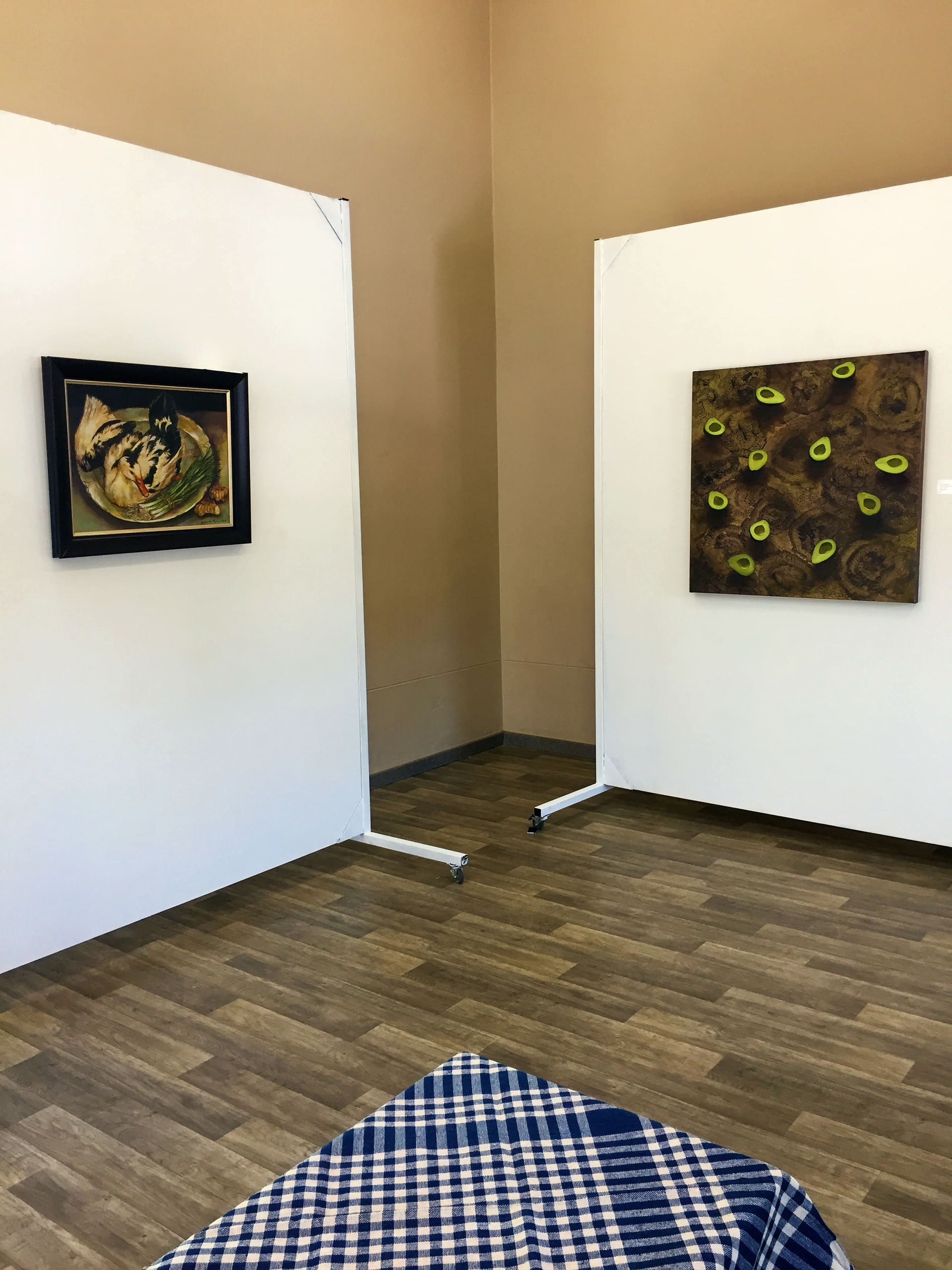 A small art gallery room with light brown walls and wooden floors. Two artworks hang on white partition walls: one is a framed abstract painting with animals; the other is a square piece featuring green and yellow circles. A checked cloth covers a surface in the foreground.