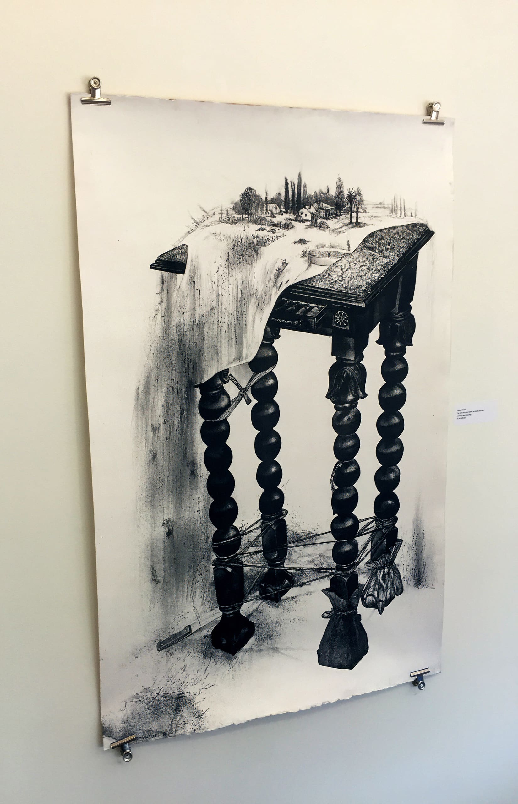 A realistic pencil drawing features a wooden table with ornate, spiralled legs. The tabletop melts and drips like cloth, blending into a landscape of trees and small houses. The artwork is pinned to a white wall with metal clips.