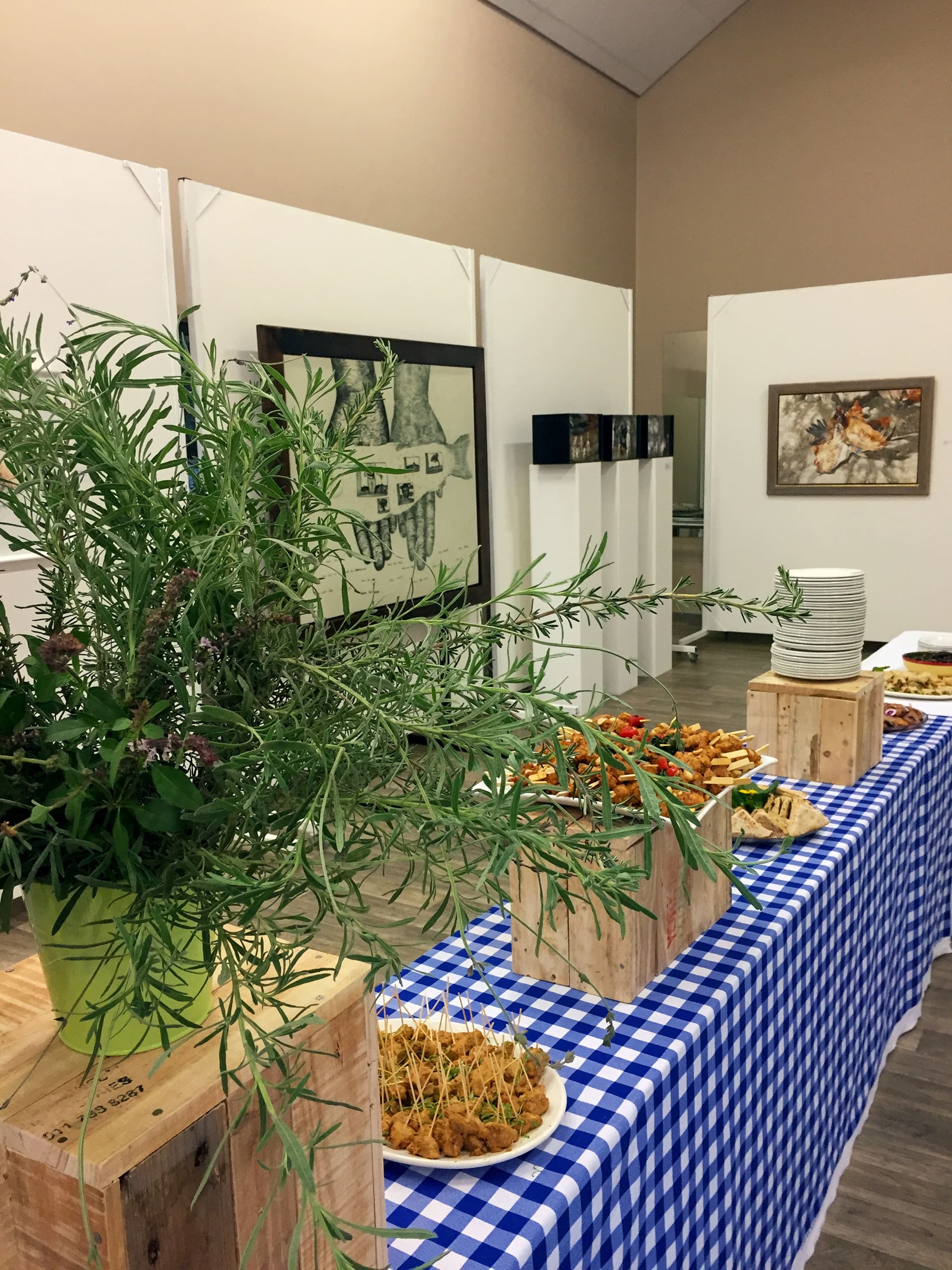A buffet table with a blue and white chequered cloth holds plates of appetisers and stacks of white plates. Wooden crates are used for display, and a large potted plant decorates the table. Framed art hangs on beige and white walls in the background.