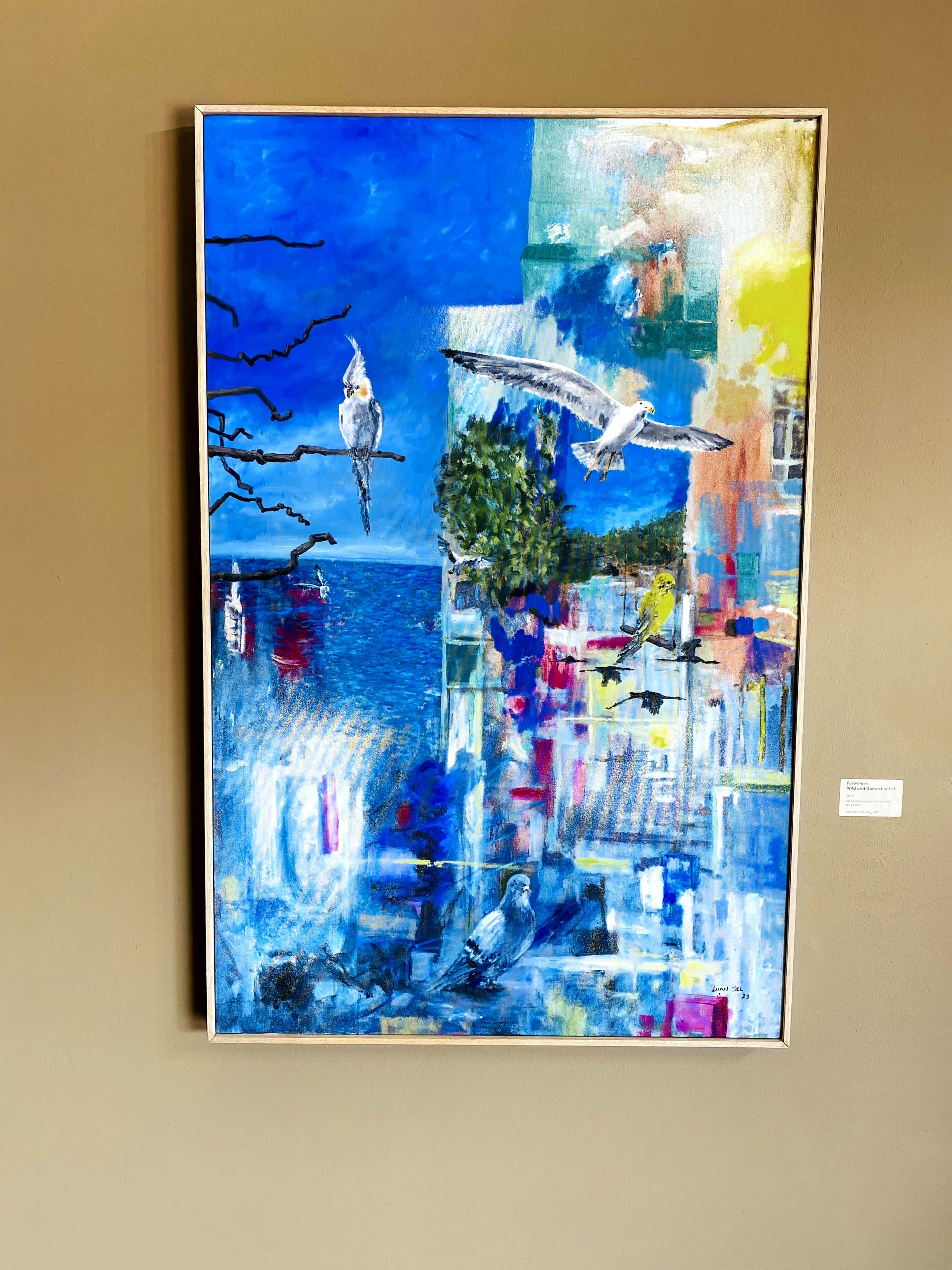 A colourful painting on a tan wall depicts three cockatiels, abstract cityscapes, blue sky, ocean, tree, geometric shapes, silhouetted tree branches, and vibrant brushstrokes blending urban and natural environments. A small label is mounted beside the artwork.