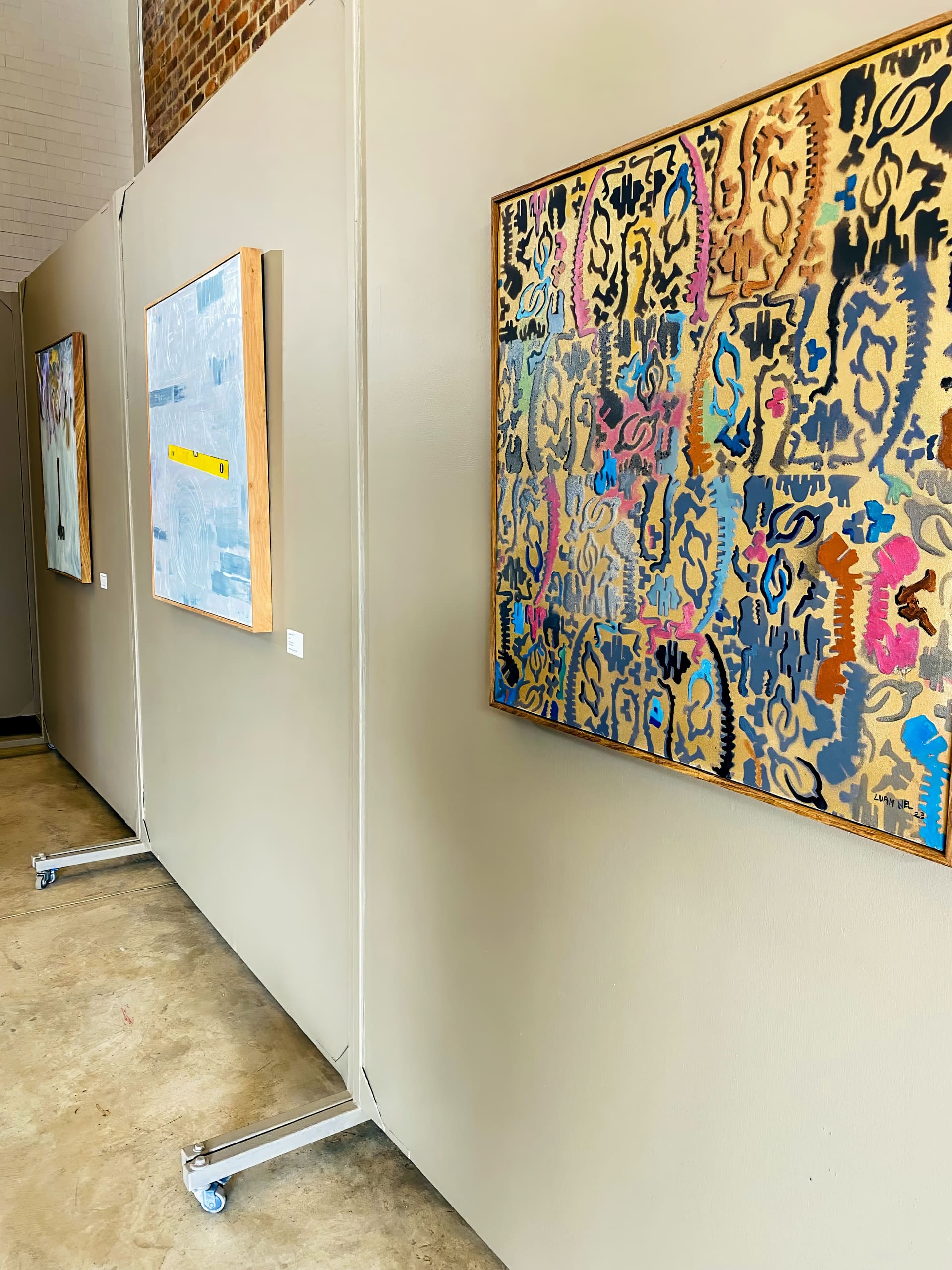 Three abstract paintings hang on beige partition walls in an art gallery. The closest painting features colourful, intricate patterns. The other two artworks, further down the wall, appear more muted and geometric. The floor is concrete, and part of a brick wall is visible.